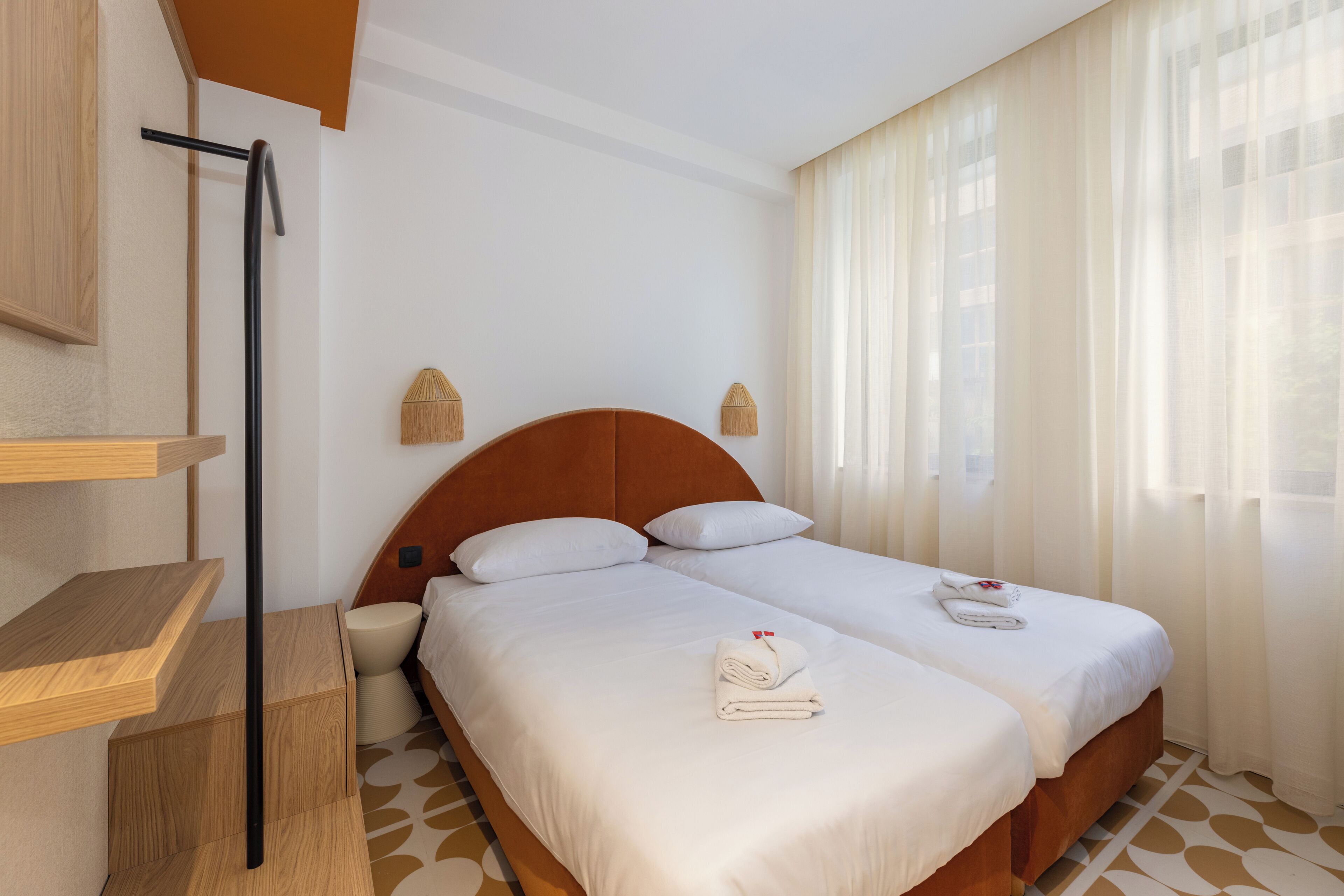 Une chambre d’hôtel avec un lit double, une tête de lit en bois, une table de chevet, une petite lampe de chevet et une fenêtre avec des rideaux légers.