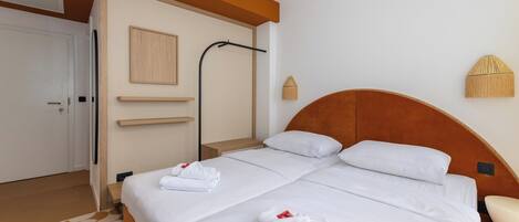 Habitación de hotel con dos camas, cabecera de madera, puerta blanca, perchero y lámpara fijada a la pared.