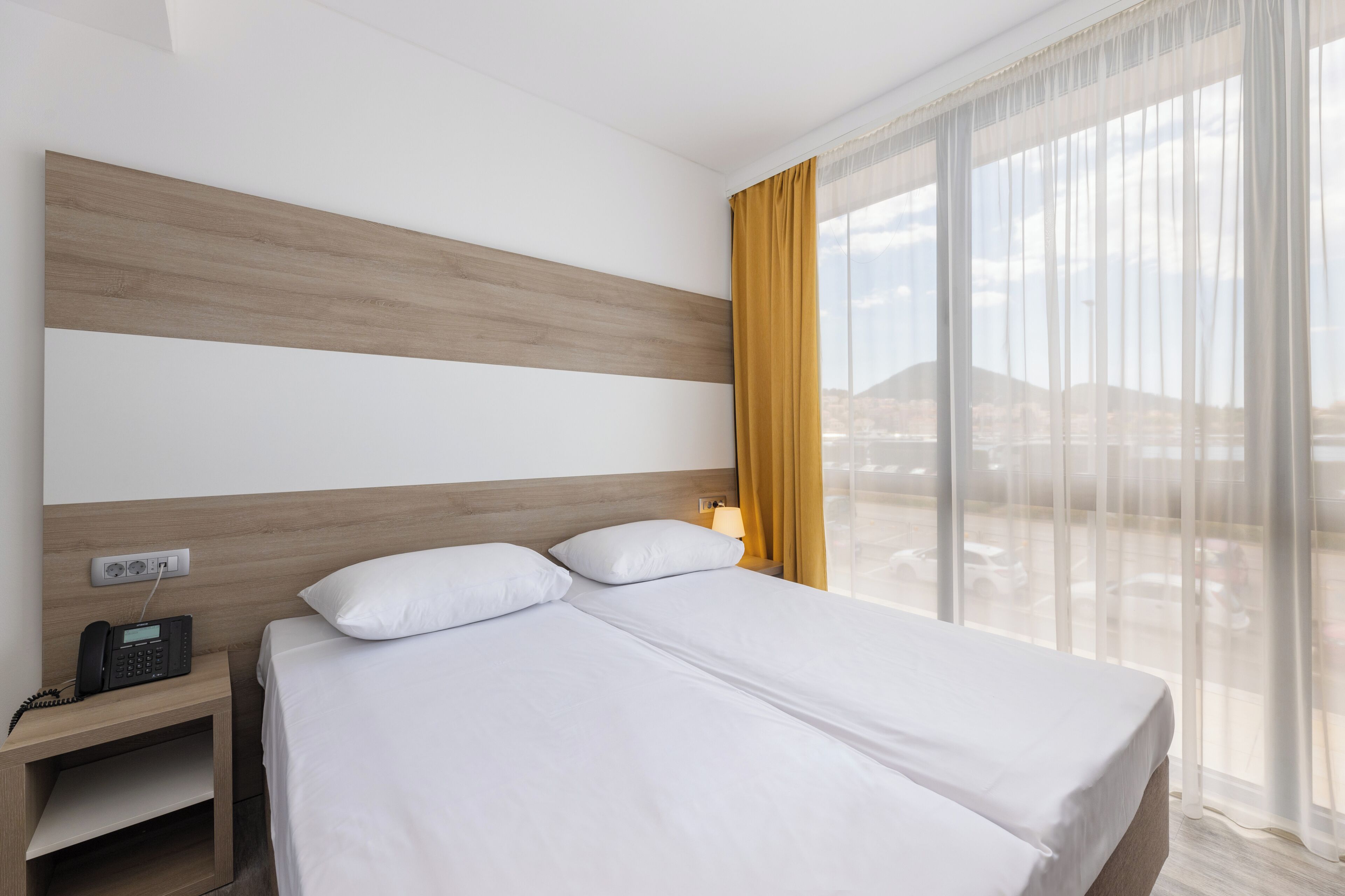 A hotel room with a bed, a bedside table with a phone, a lamp, and a view of a cityscape through large windows.