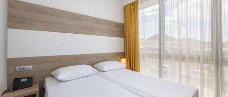 A hotel room with a bed, a bedside table with a phone, a lamp, and a view of a cityscape through large windows.