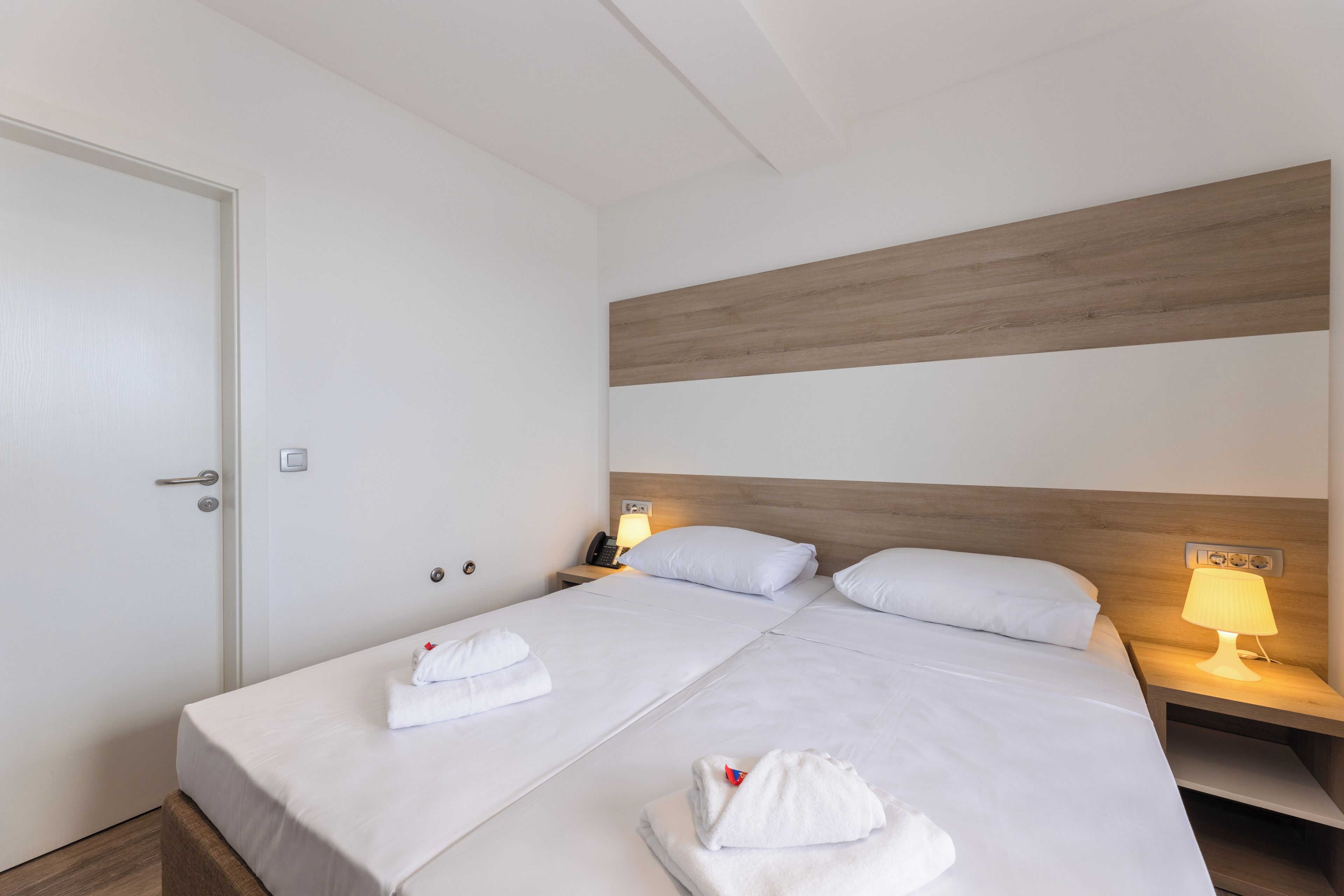 Quarto de hotel com duas camas, cabeceira de madeira, abajures e uma porta.