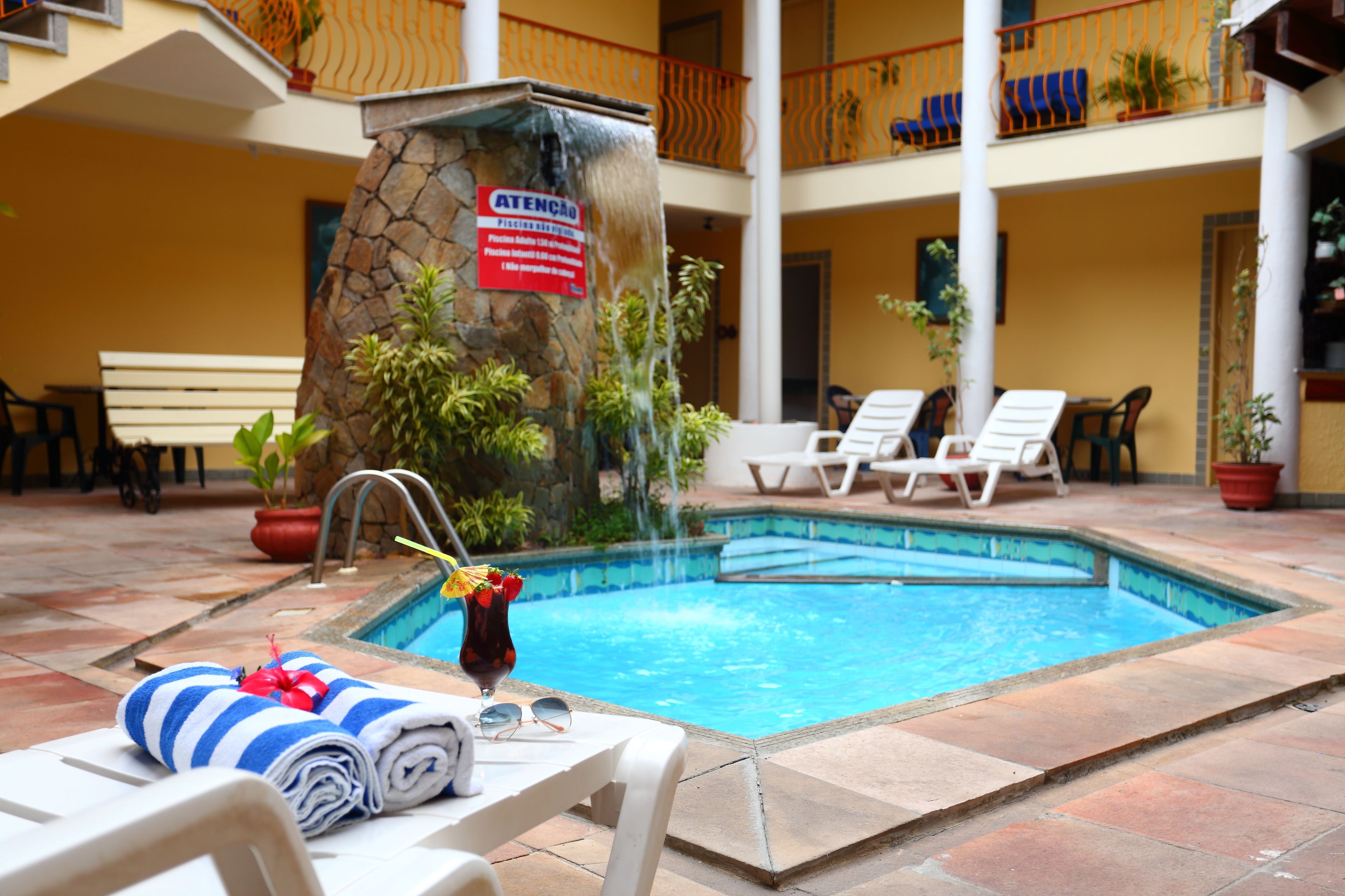 A pool area with lounge chairs, a stone wall with a waterfall, and a sign in Portuguese.