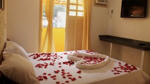 A hotel room with a bed covered in rose petals, a television on the wall, and a window with yellow curtains. - Marlim Porto Hotel (Porto Seguro)