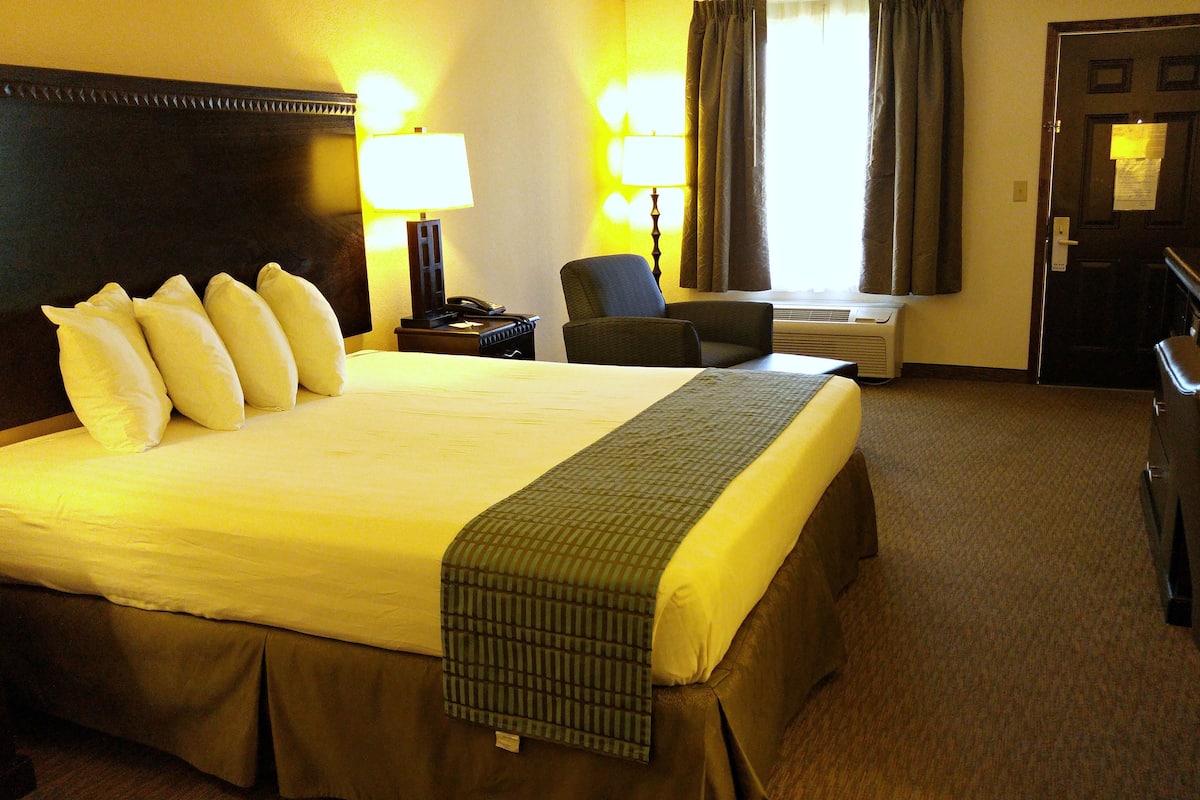 A hotel room with a large bed, two bedside lamps, a chair, a desk with a computer, and a door with a sign.