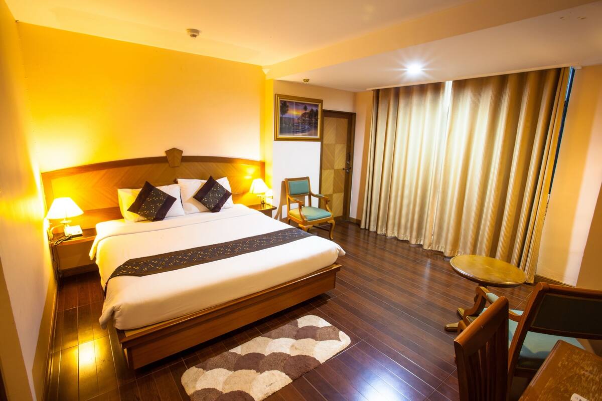 Een hotelkamer met een groot bed, twee bedlampen, een stoel, een kleine tafel en uitzicht buiten door gordijnen.