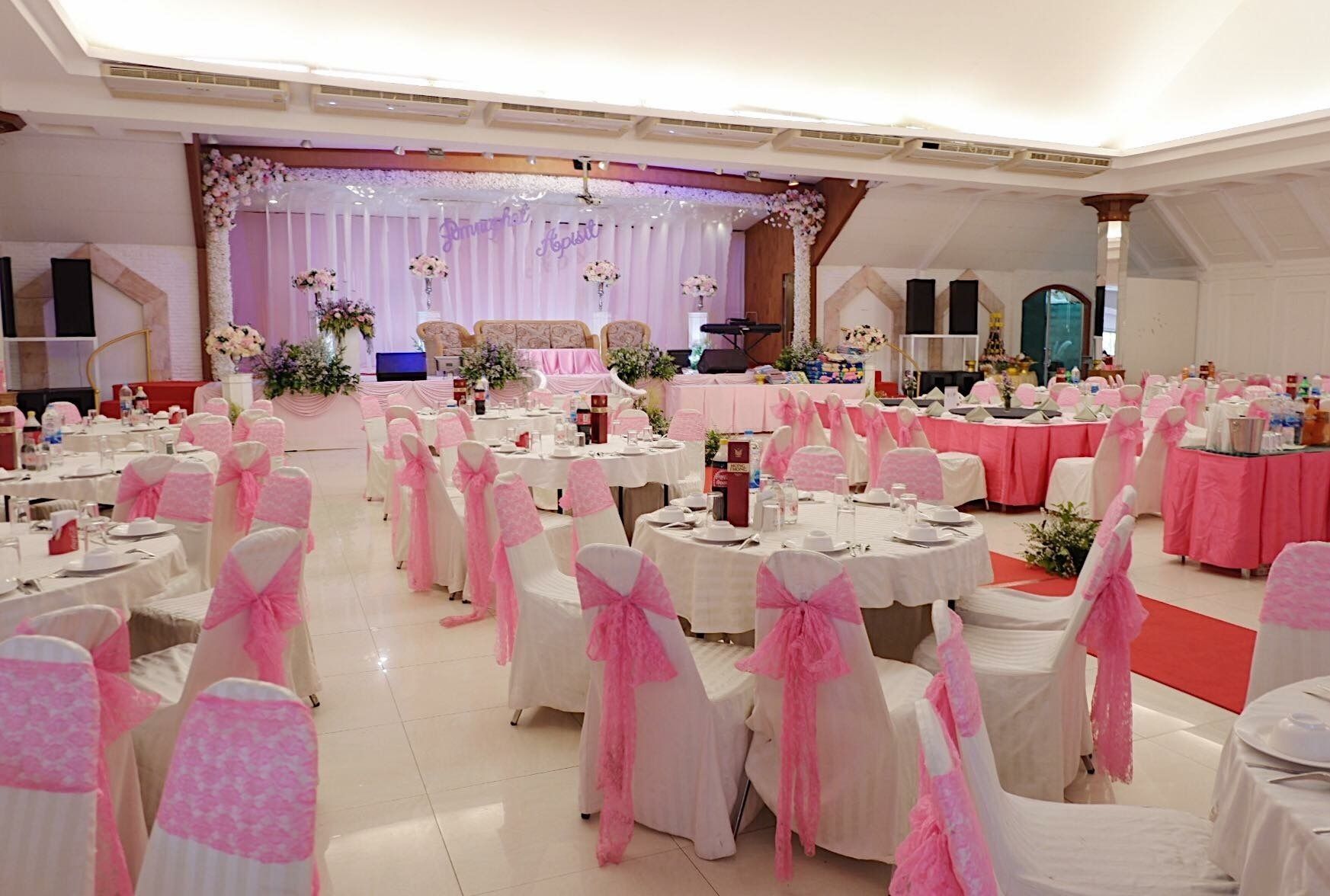 A banquet hall with round tables, white and pink chairs, and a stage with floral decorations.