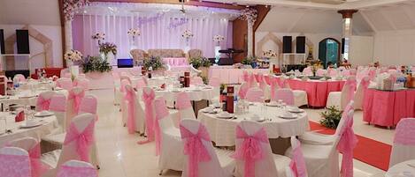 A banquet hall with round tables, white and pink chairs, and a stage with floral decorations.