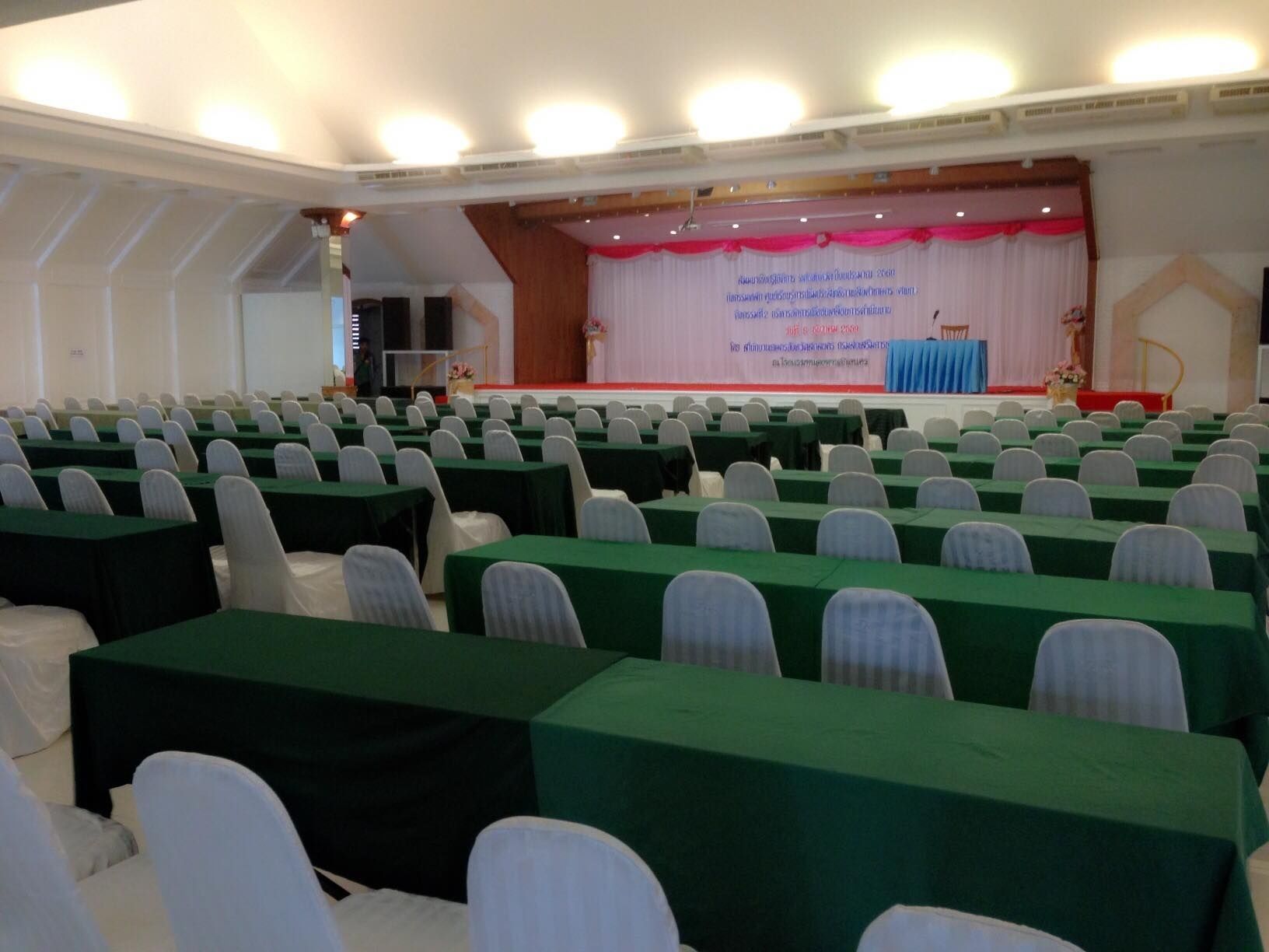 Un salón de conferencias con filas de mesas y sillas, un escenario con un atril, y un cartel.