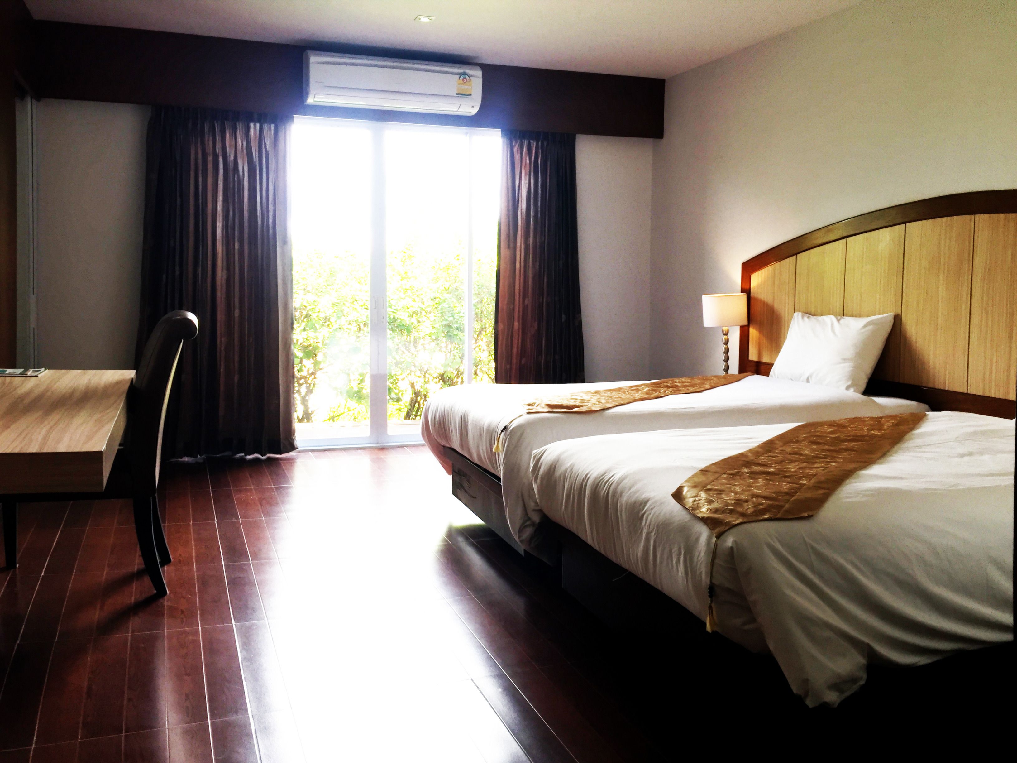 A hotel room with a bed, a desk, and a chair. There is an air conditioning unit above the bed and a window with curtains.