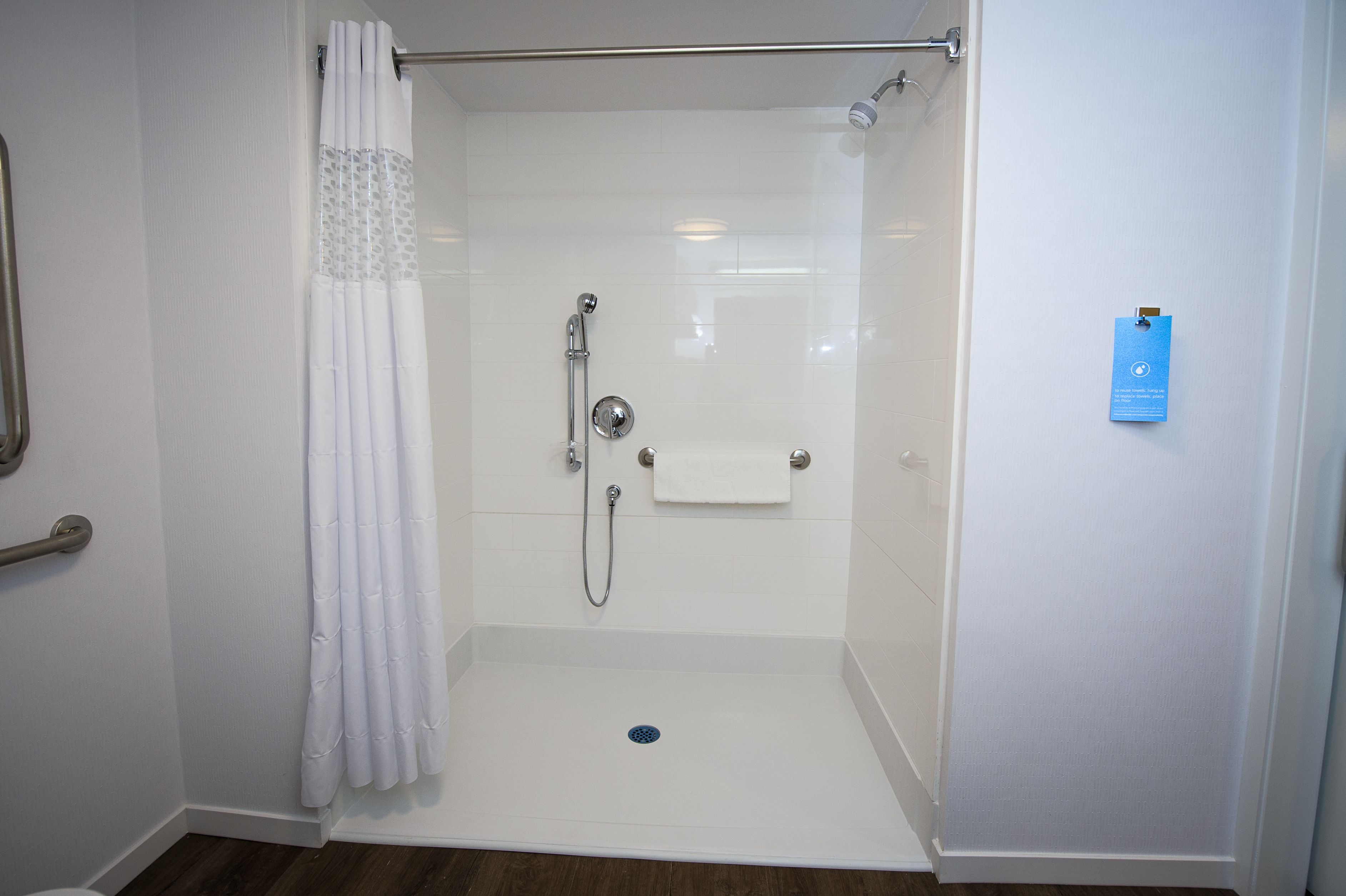 Une douche à l’italienne avec un rideau blanc, une douchette à main, un porte-serviettes et une carte d’information bleue accrochée au mur.