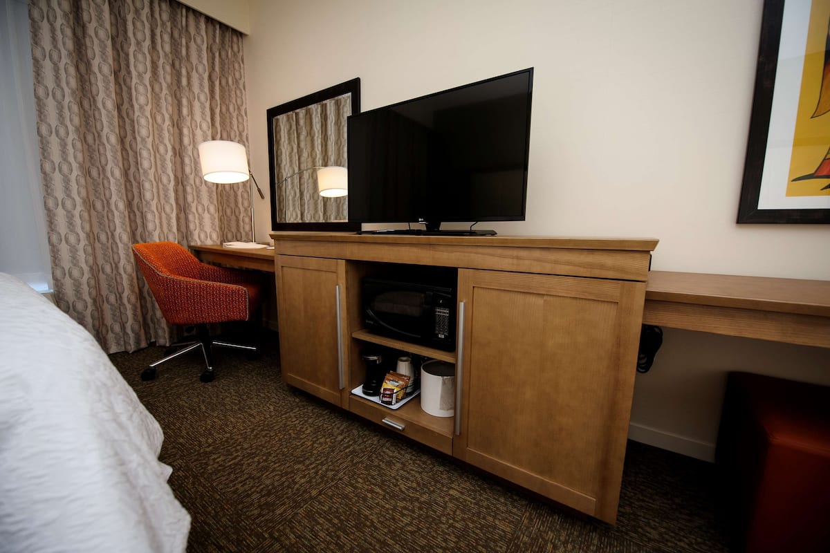 A hotel room with a bed, a desk with a TV, a chair, and a minibar.