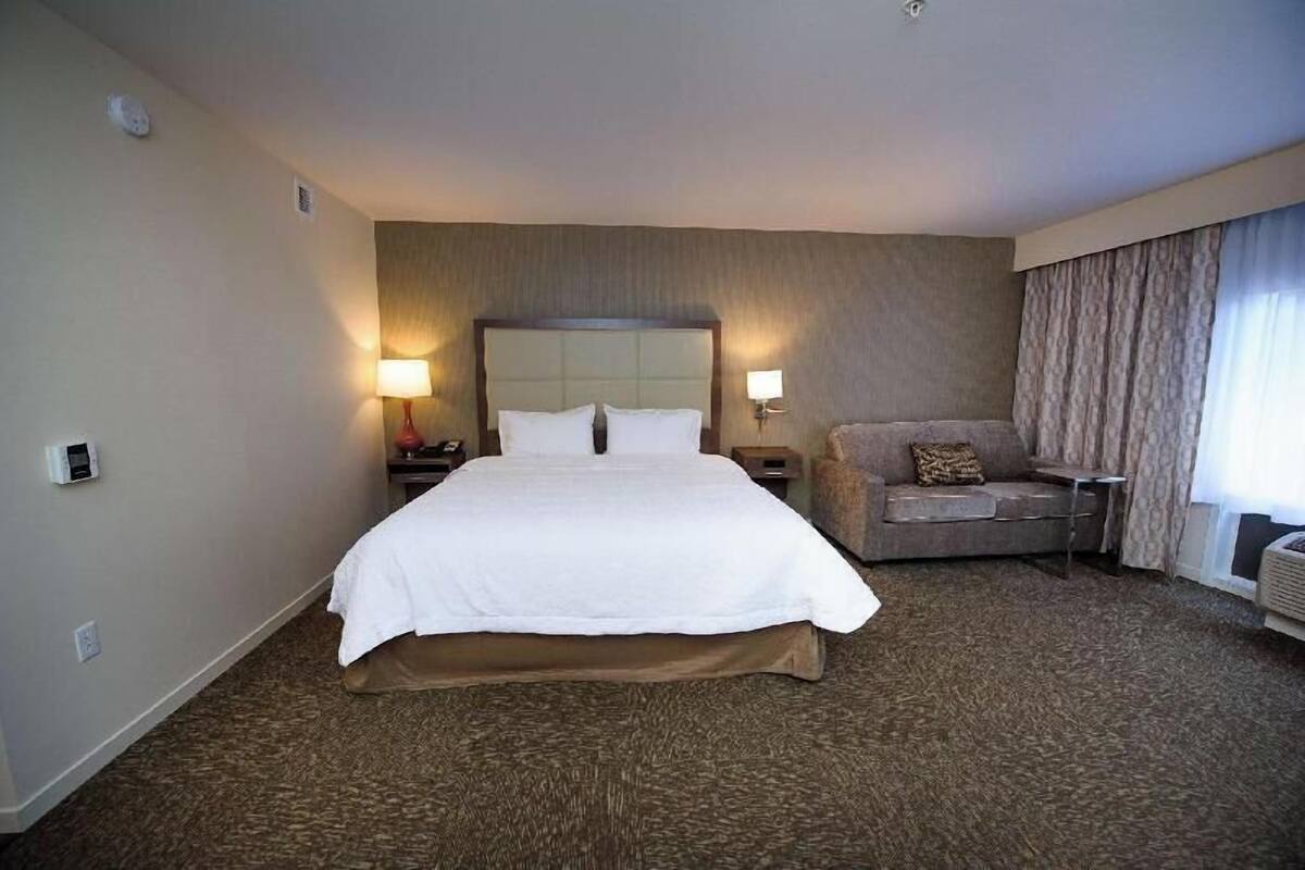 A hotel room with a large bed, a sofa, a nightstand with a lamp, and a window with curtains.