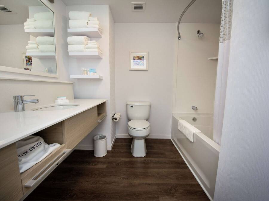 A modern bathroom with a white sink, toilet, and shower. There are shelves with towels and a trash can.