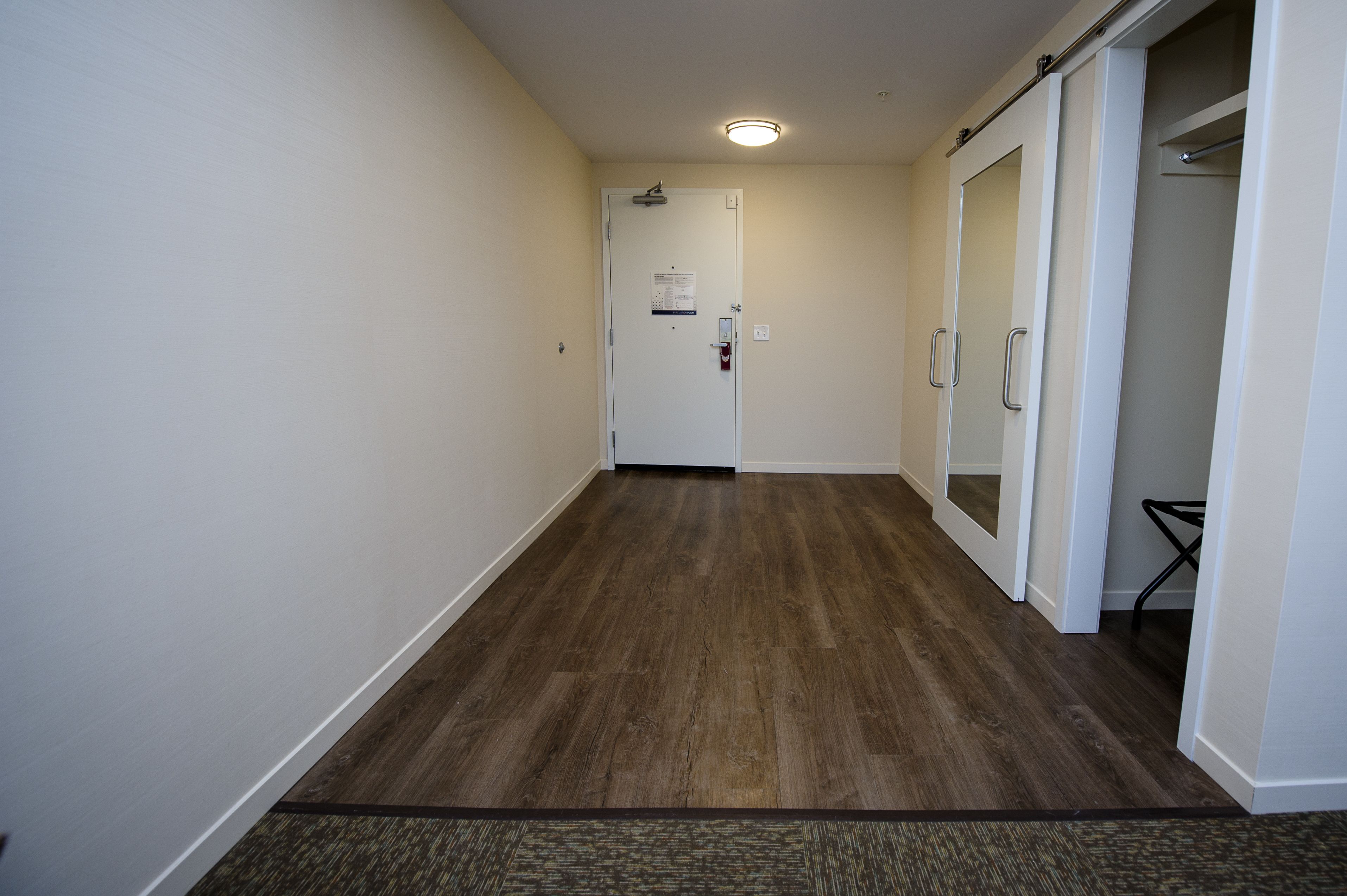 A hallway with a wooden floor, white walls, and a door with a fire alarm.