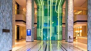 Eine moderne Hotelhalle mit einer zentralen Glaswand, die mit vertikalen Lichtleisten, Marmorsäulen und einer digitalen Anzeige versehen ist.