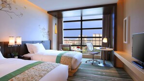 Ein Hotelzimmer mit zwei Betten, einem Schreibtisch, einem Sessel, einem Fernseher und einem großen Fenster mit Blick auf die Stadt.