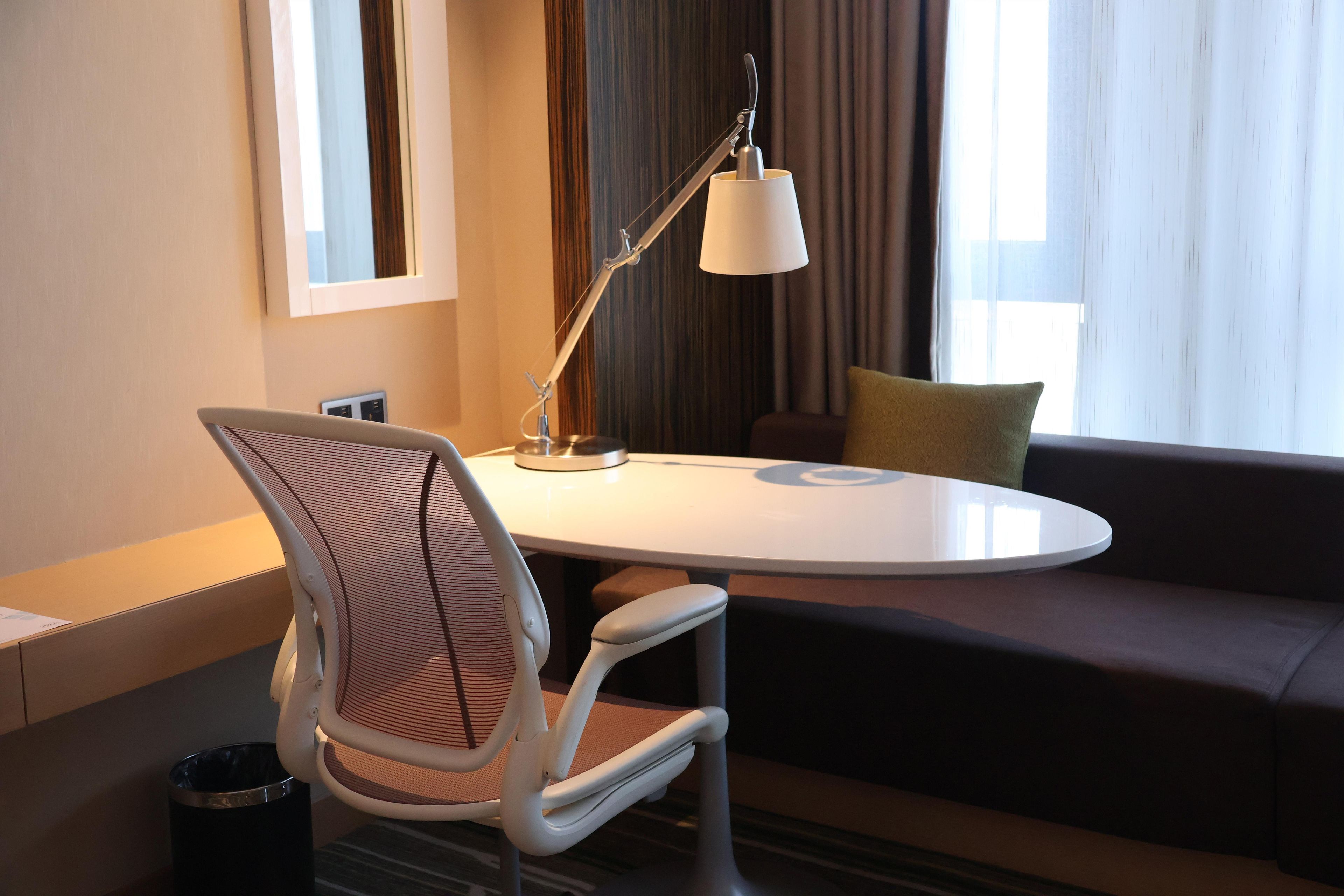 Una habitación de hotel moderna con una mesa redonda, una silla blanca, un sofá, una lámpara y una ventana con cortinas.