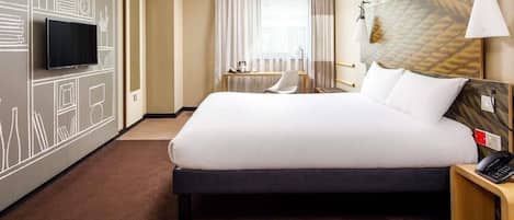 A hotel room with a large bed, a TV mounted on the wall, a bedside table with a lamp, and a window with curtains.