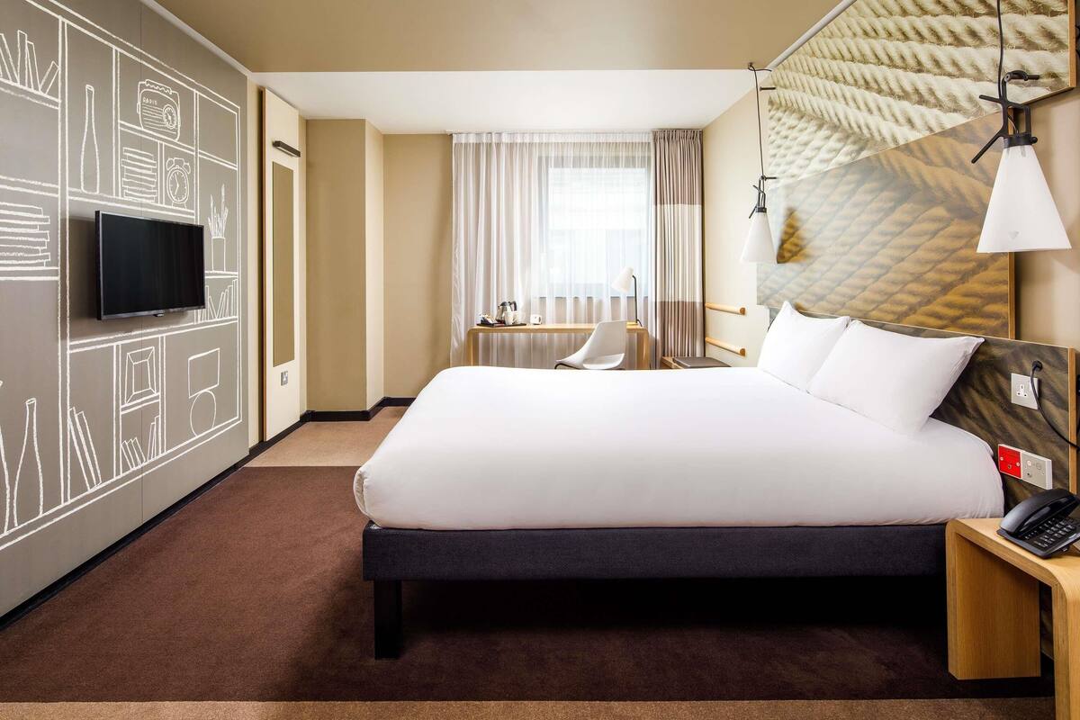 A hotel room with a large bed, a flat-screen TV mounted on the wall, a desk with a chair, and a window with curtains.