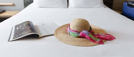A neatly made bed with white linens, a straw hat with a colorful scarf, and an open magazine on the bed.