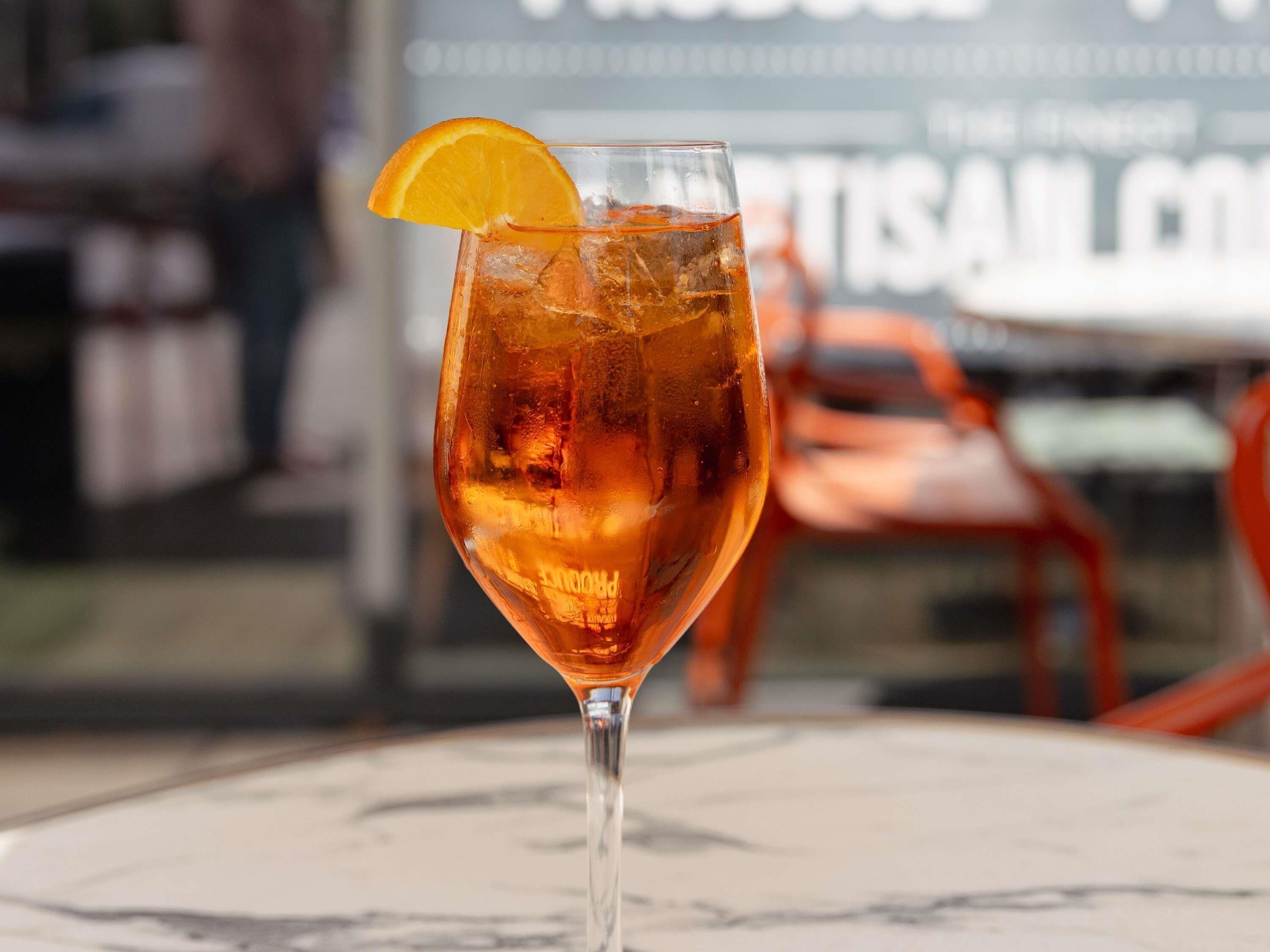 A glass of amber-colored drink with ice cubes and an orange slice garnish on a table.