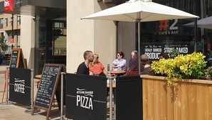 An outdoor seating area of a cafe with a sign for stone-baked pizza and a sign for a coffee shop. - ibis Cambridge Central Station (Cambridge)