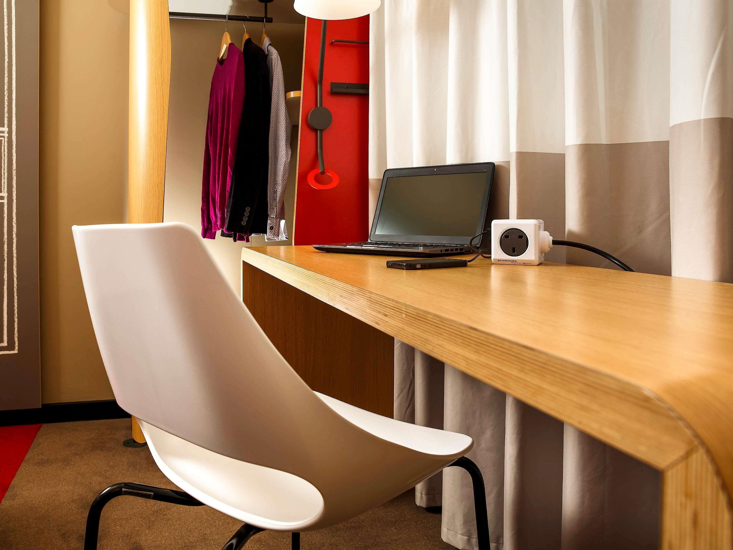 A hotel room with a wooden desk, a laptop, a power strip, a white chair, and a coat rack with hanging clothes.