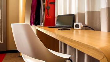 A hotel room with a wooden desk, a laptop, a power strip, a white chair, and a coat rack with hanging clothes.