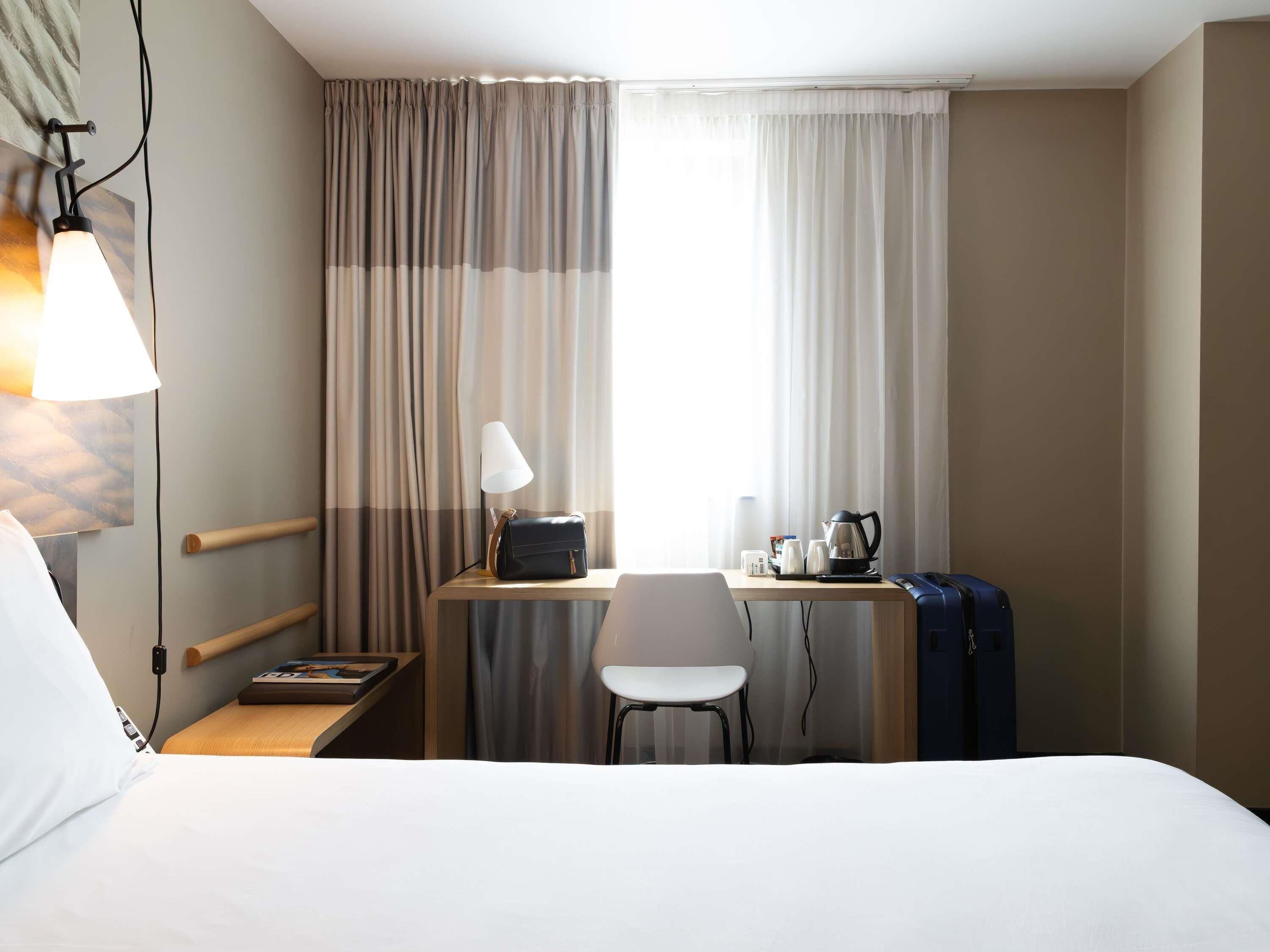 A hotel room with a bed, desk, chair, lamp, and a window with curtains.