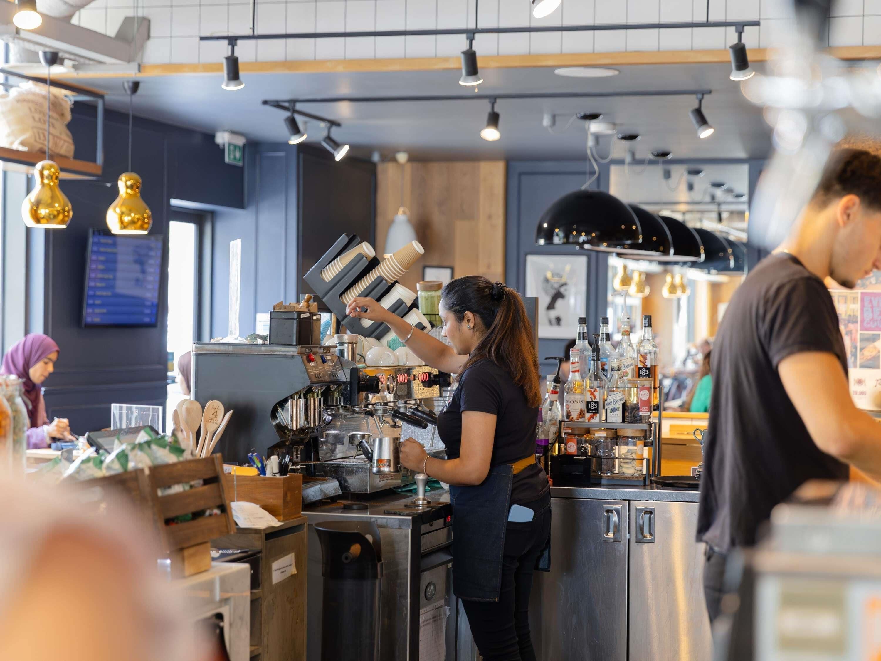 Interno di una caffetteria con un barista che prepara bevande, una lavagna con il menu e diverse attrezzature per la preparazione del caffè.