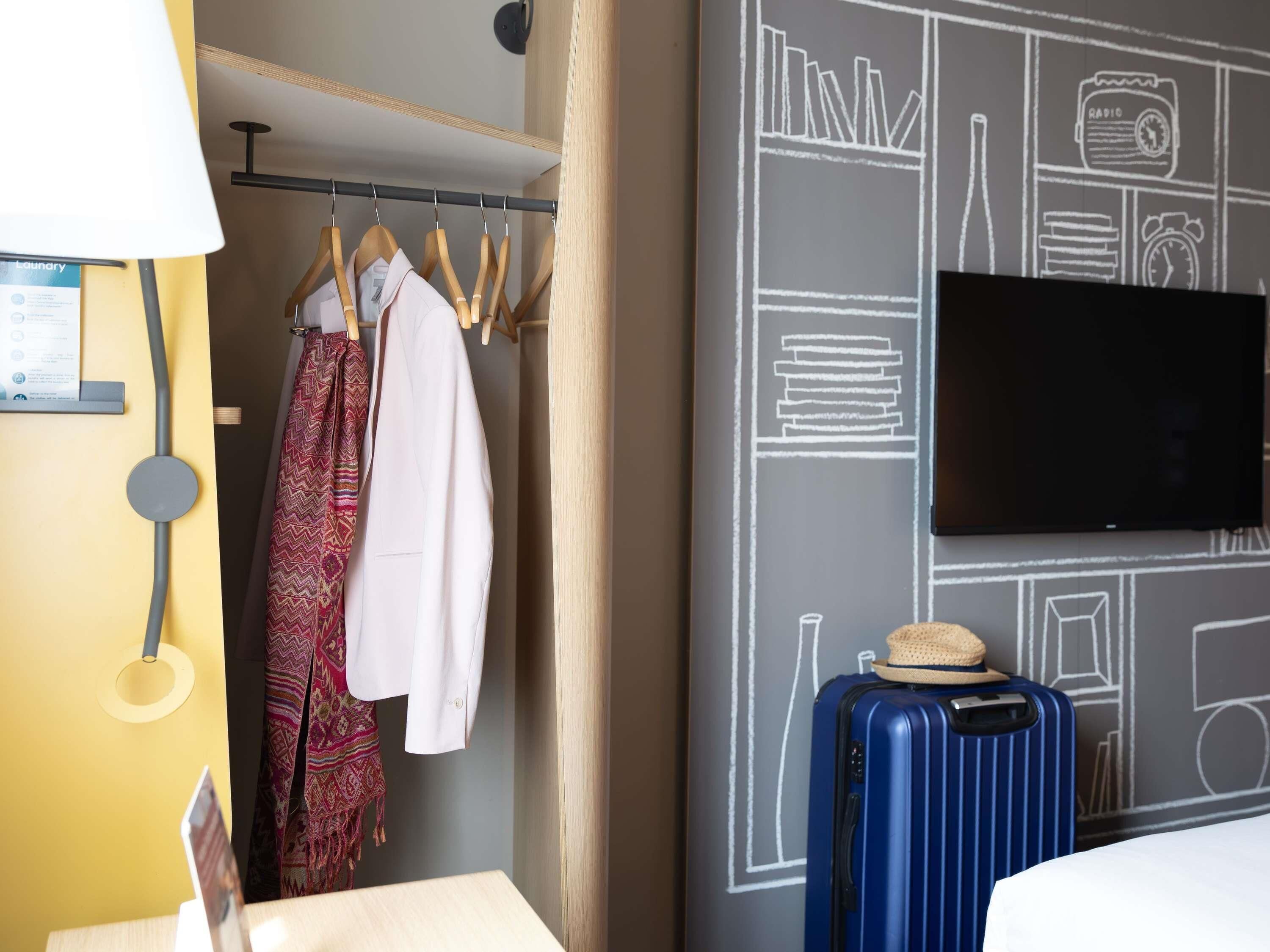 A hotel room with a bed, a wardrobe with clothes, a suitcase, and a wall with a chalkboard design.