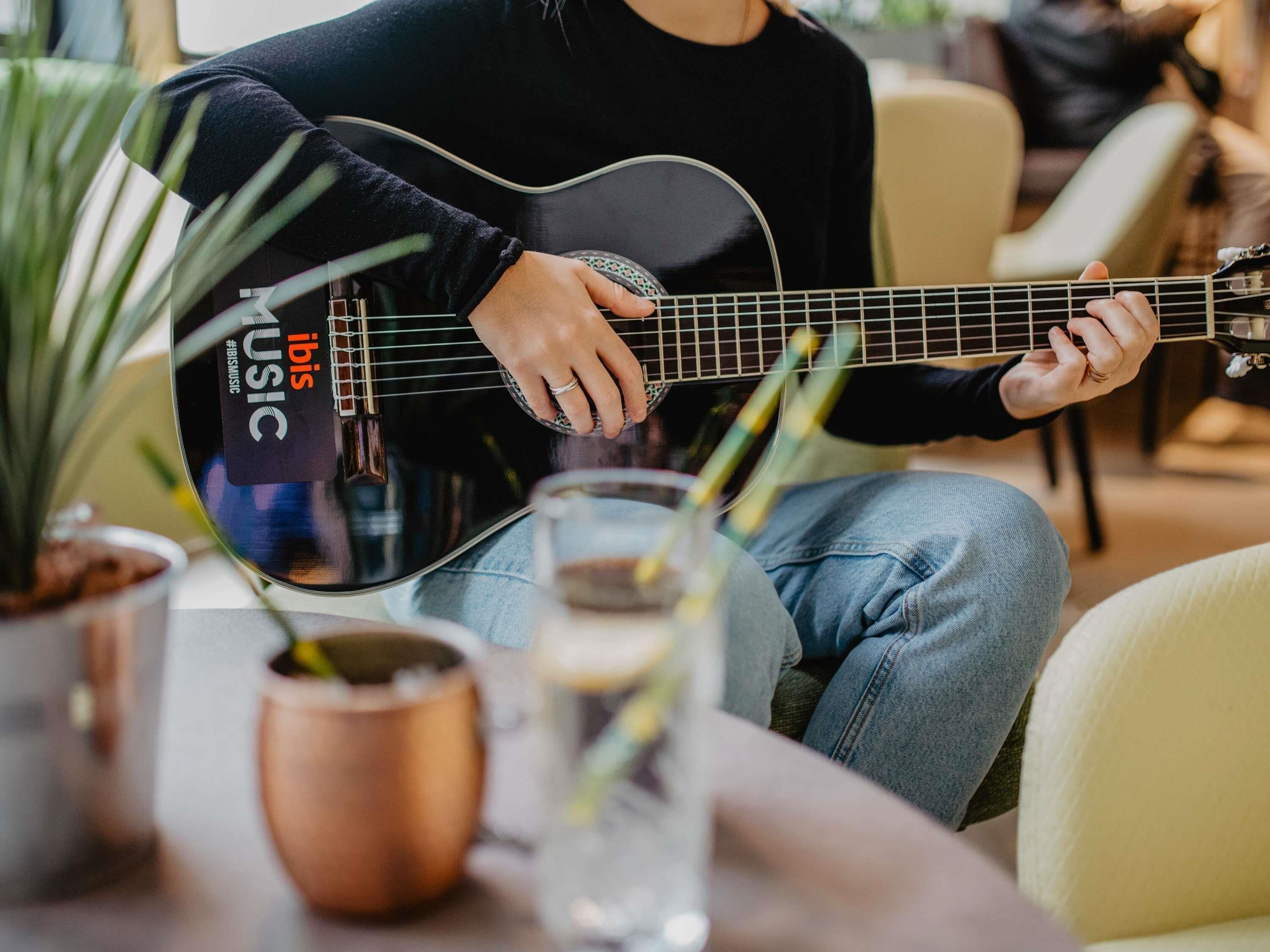 Una persona suona la chitarra in un ambiente interno accogliente, con un vaso di piante e un bicchiere d'acqua sul tavolo.