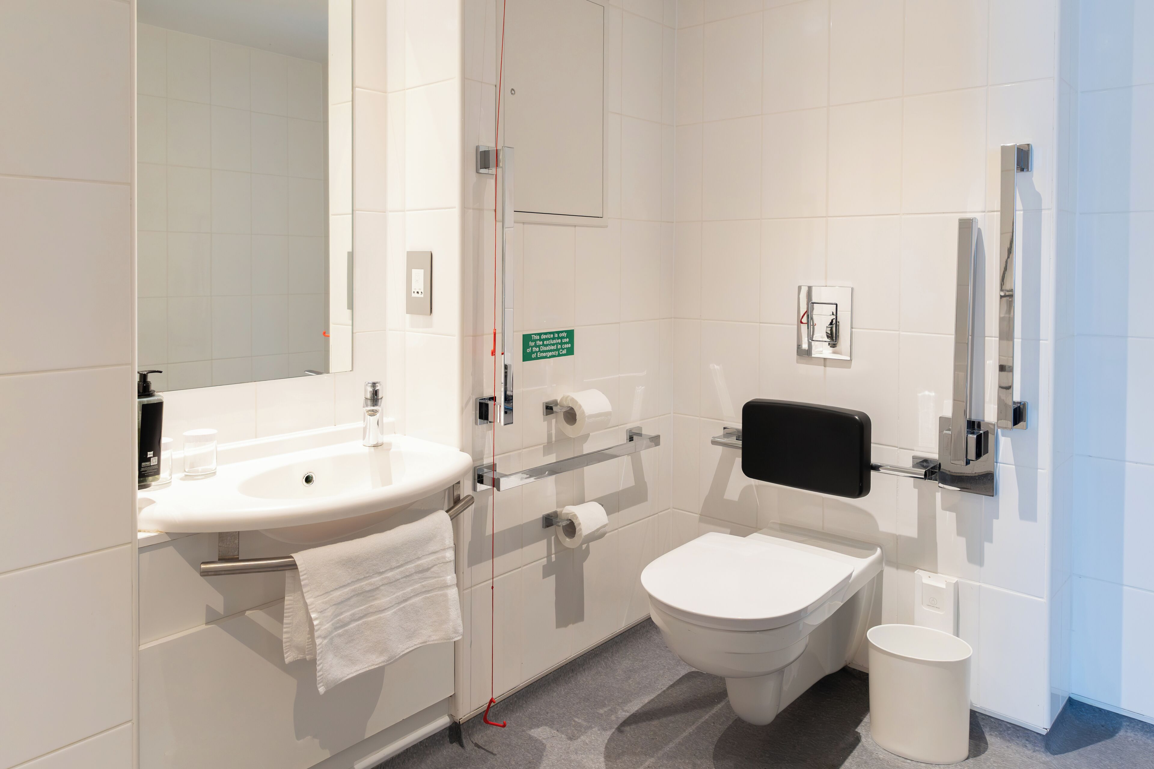 Un baño moderno con lavamanos, inodoro y bidé blancos, equipado con un perchero y un cubo de basura.