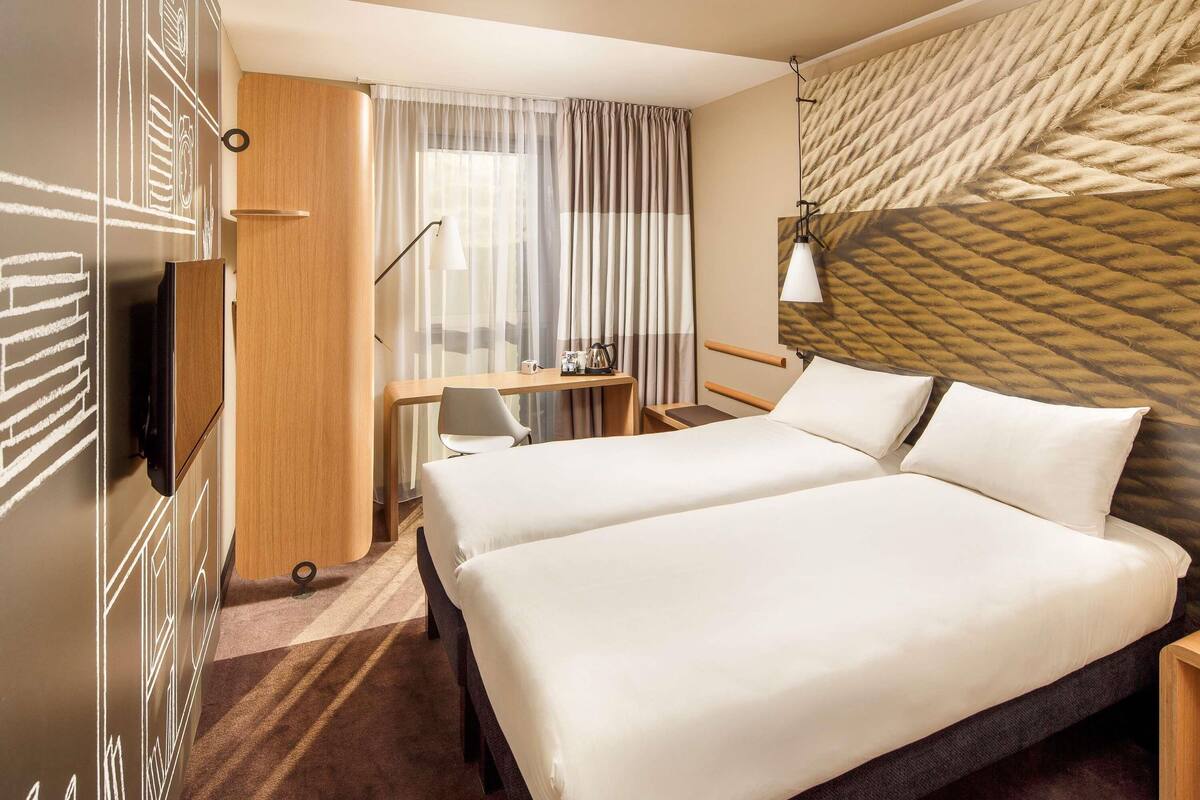 A hotel room with two beds, a desk, a chair, a TV, and a patterned wall.