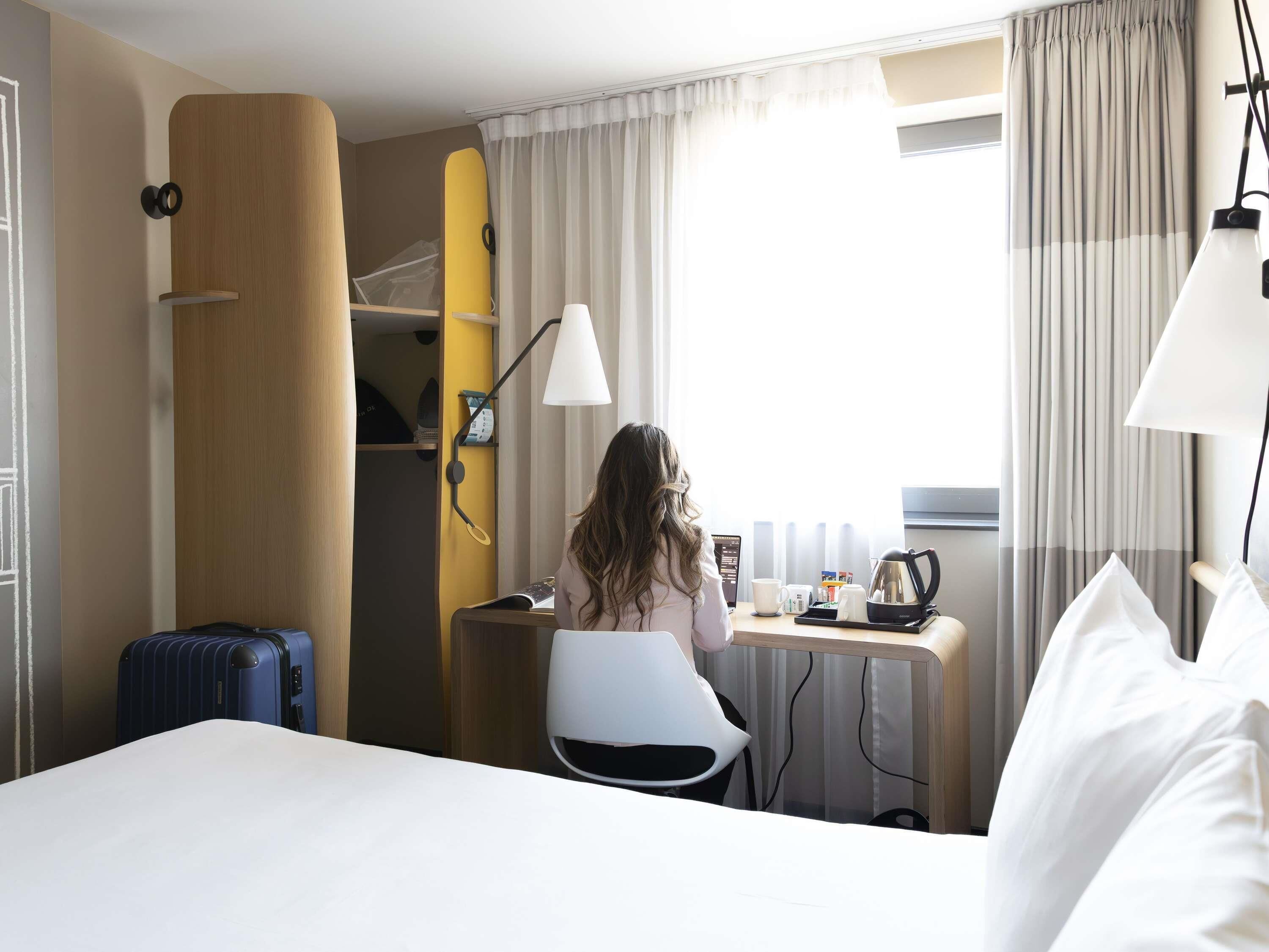 Hotellihuone, jossa on sänky, työpöytä kannettavalla tietokoneella, tuoli, matkalaukku, surffilauta ja ikkuna, jossa on verhot.