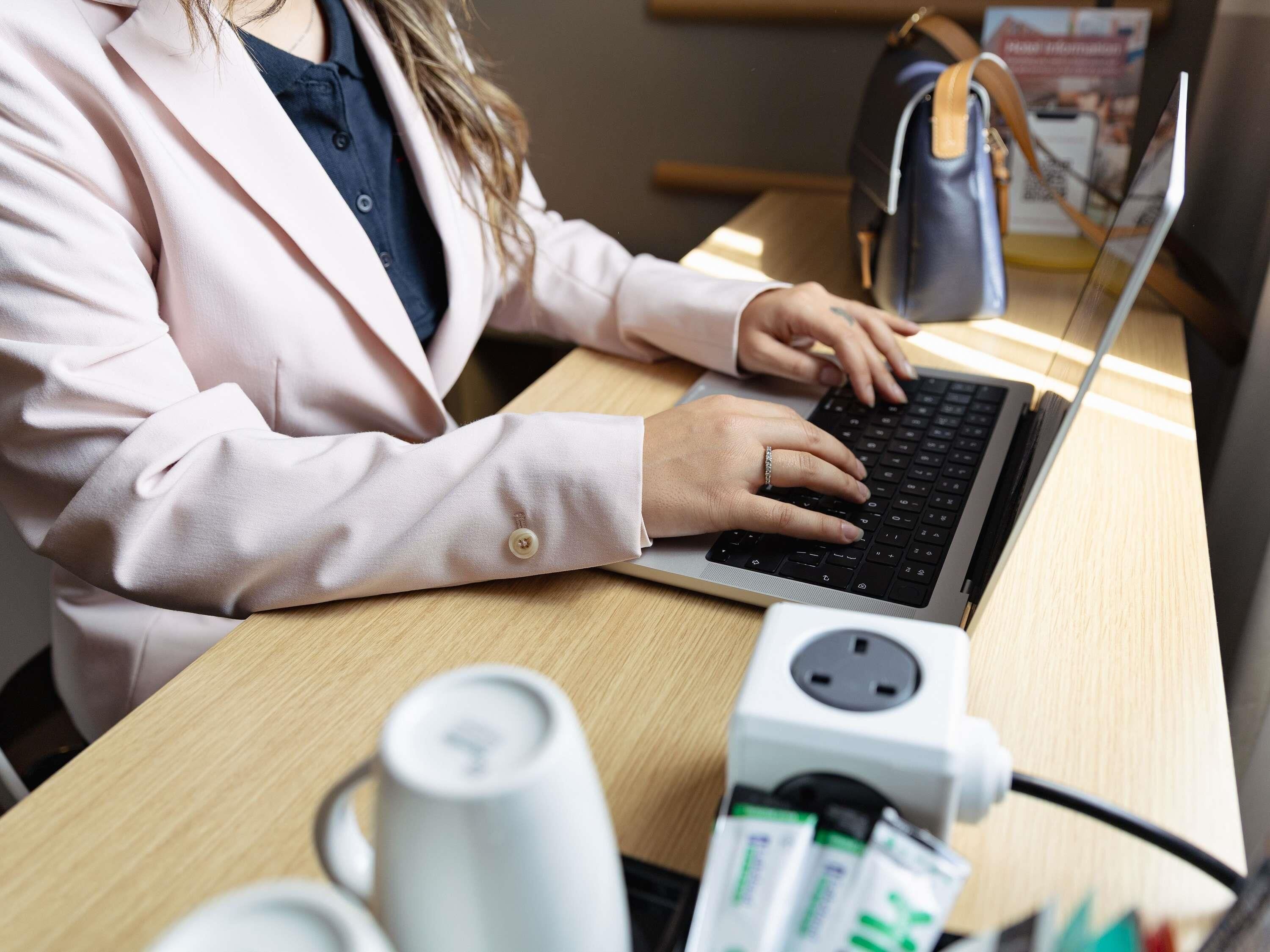 Een persoon gebruikt een laptop aan een houten tafel met een stekkerdoos en een witte mok.
