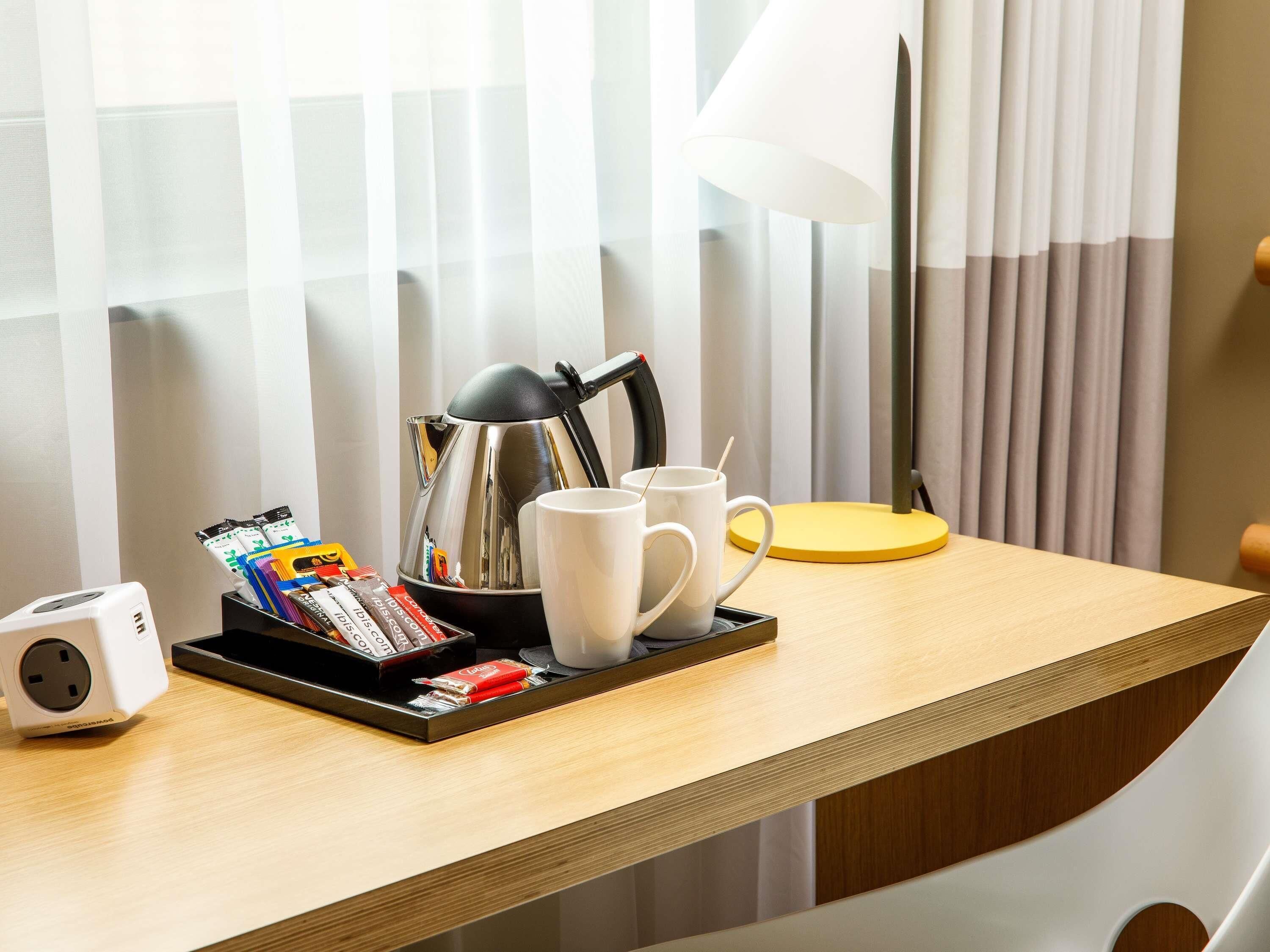 A hotel room with a wooden desk, a white mug, a silver kettle, a yellow coaster, and a tray with magazines and snacks.
