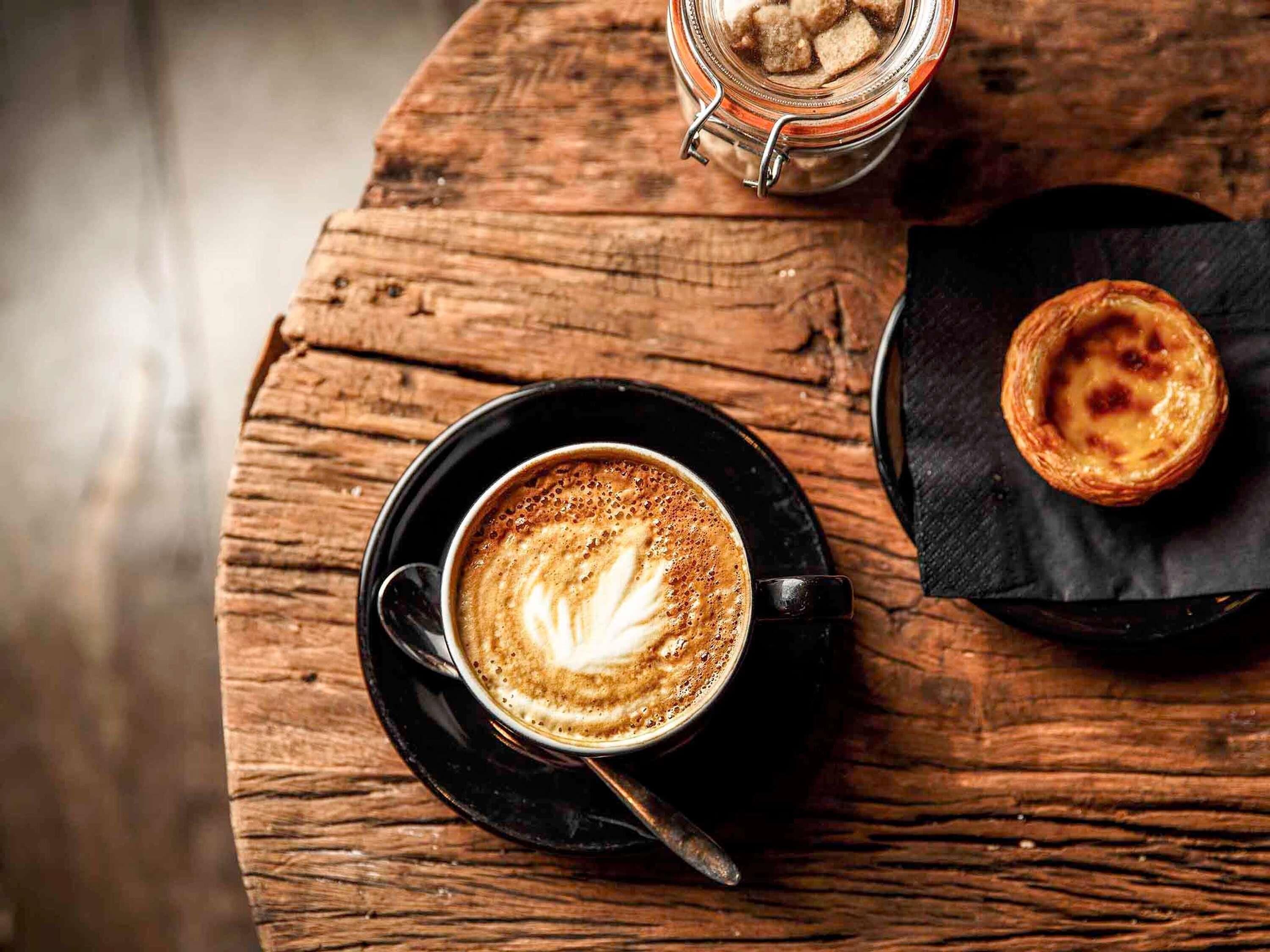 A cup of coffee with latte art on a wooden table, a jar of biscotti, and a pastry on a napkin.