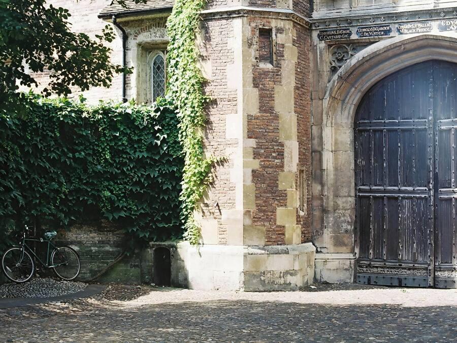 Un edificio histórico con una gran entrada en arco, una bicicleta estacionada contra la pared y un muro de ladrillo parcialmente cubierto de hiedra.