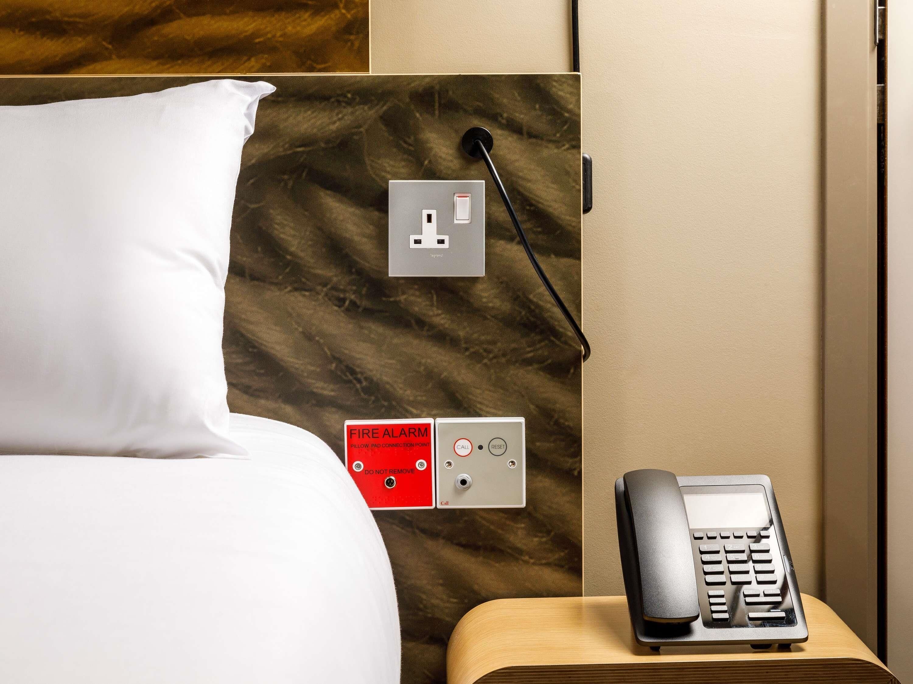 A hotel room with a bed, a bedside table with a telephone, and a wall-mounted fire alarm.