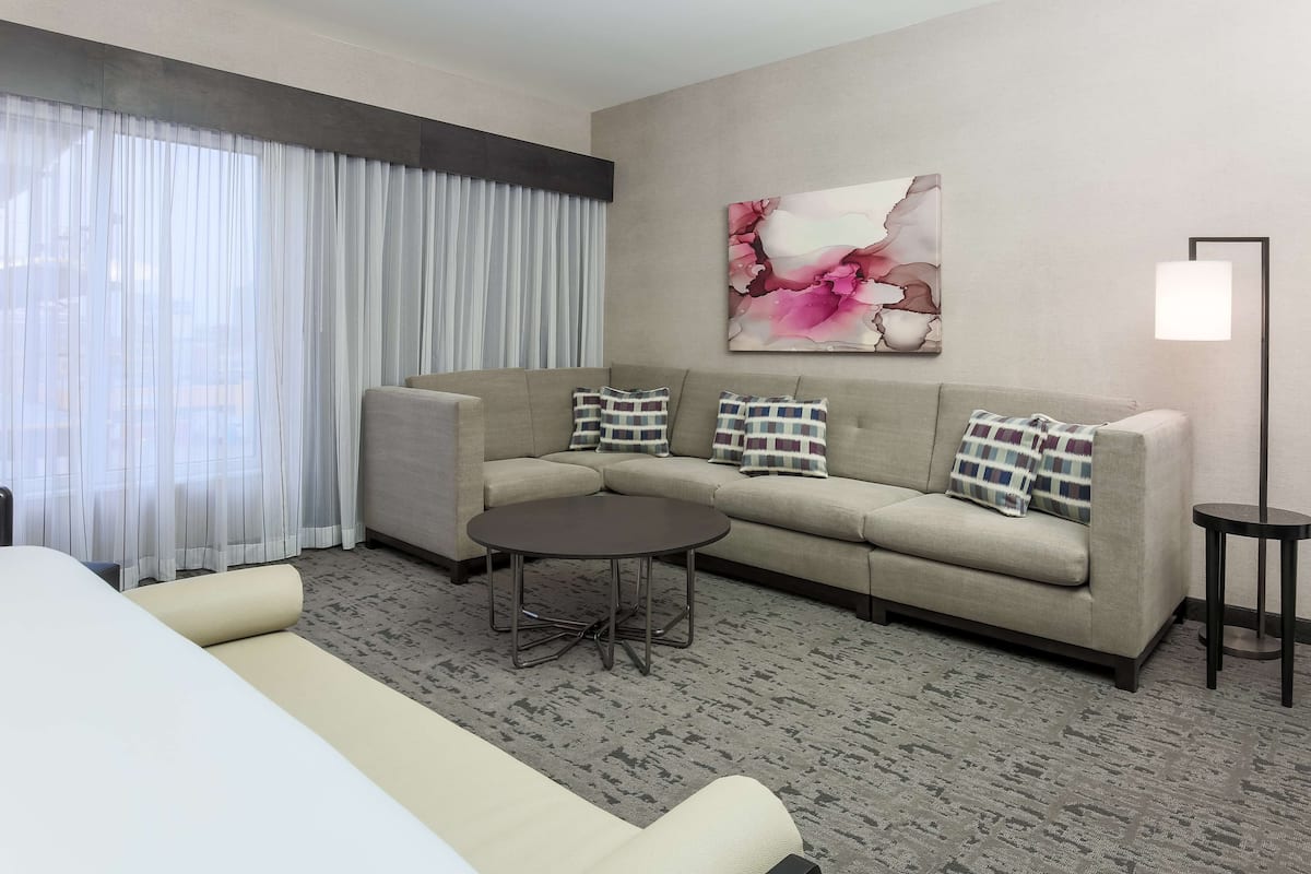 A hotel room with a beige sofa, a round coffee table, a large window with sheer curtains, and a floral artwork on the wall.