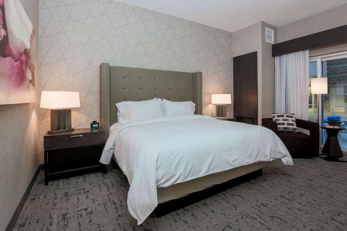 A hotel room with a large bed, two bedside tables with lamps, a chair, and a view of the outdoors.