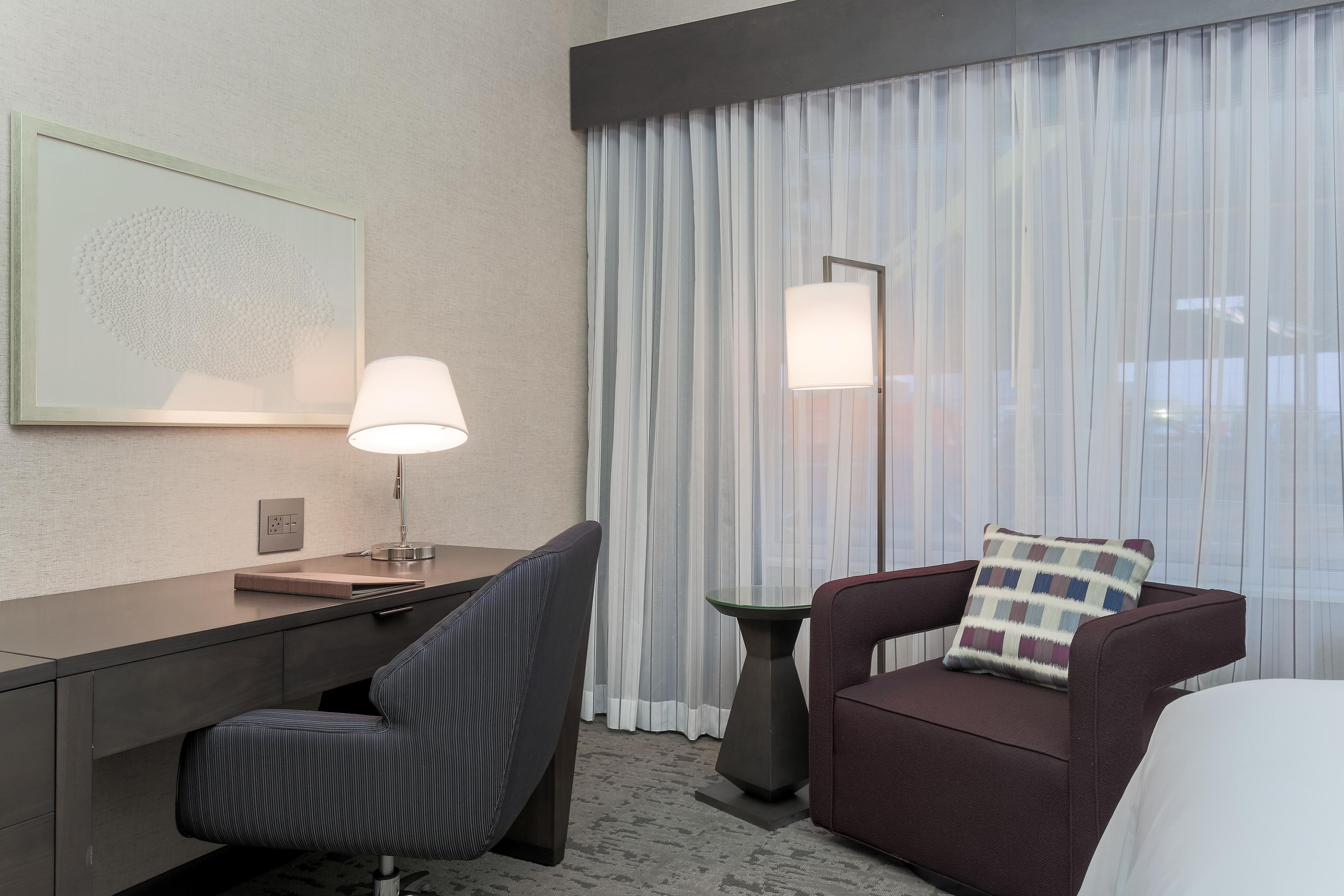 Une chambre d’hôtel avec un bureau, une chaise et un canapé avec un coussin. Il y a une lampe sur le bureau et une autre sur le sol.