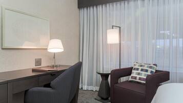 Habitación de hotel con escritorio, silla y sofá con almohada. Hay una lámpara en el escritorio y otra en el suelo.
