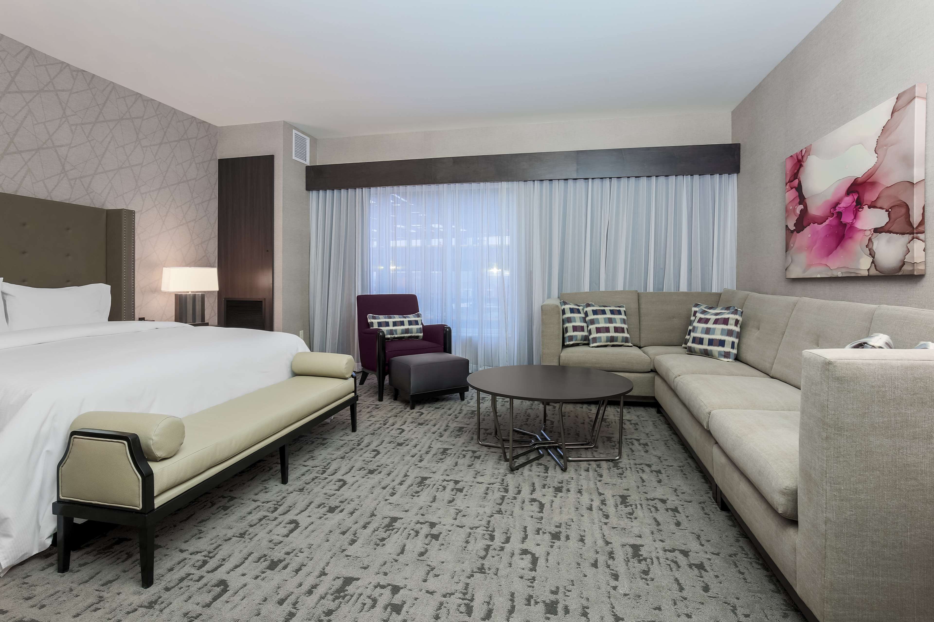 Hotelkamer met een groot bed, een bank, een kleine tafel, een stoel en een groot raam met gordijnen.