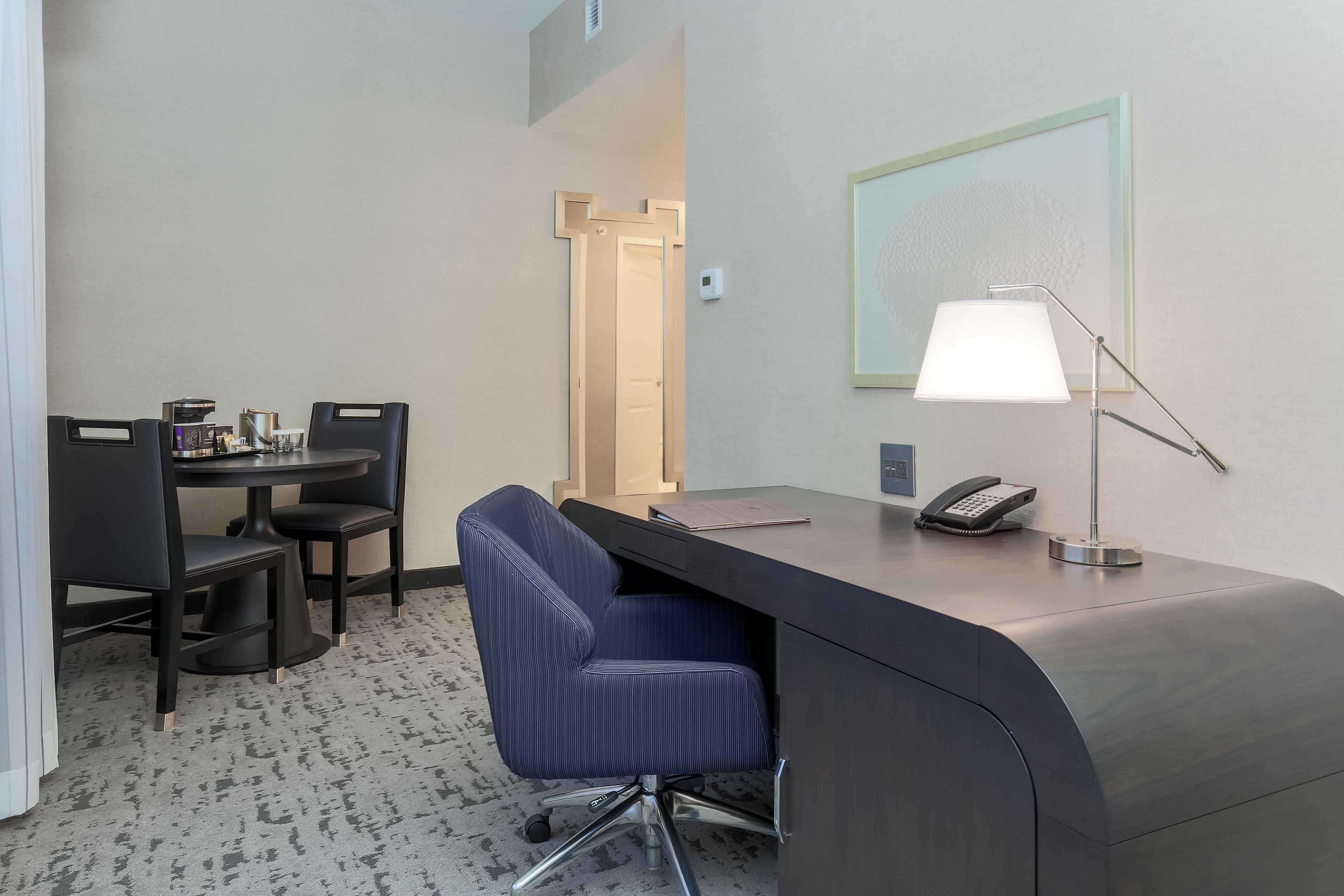 A hotel room with a desk, chairs, a lamp, a phone, and a door.