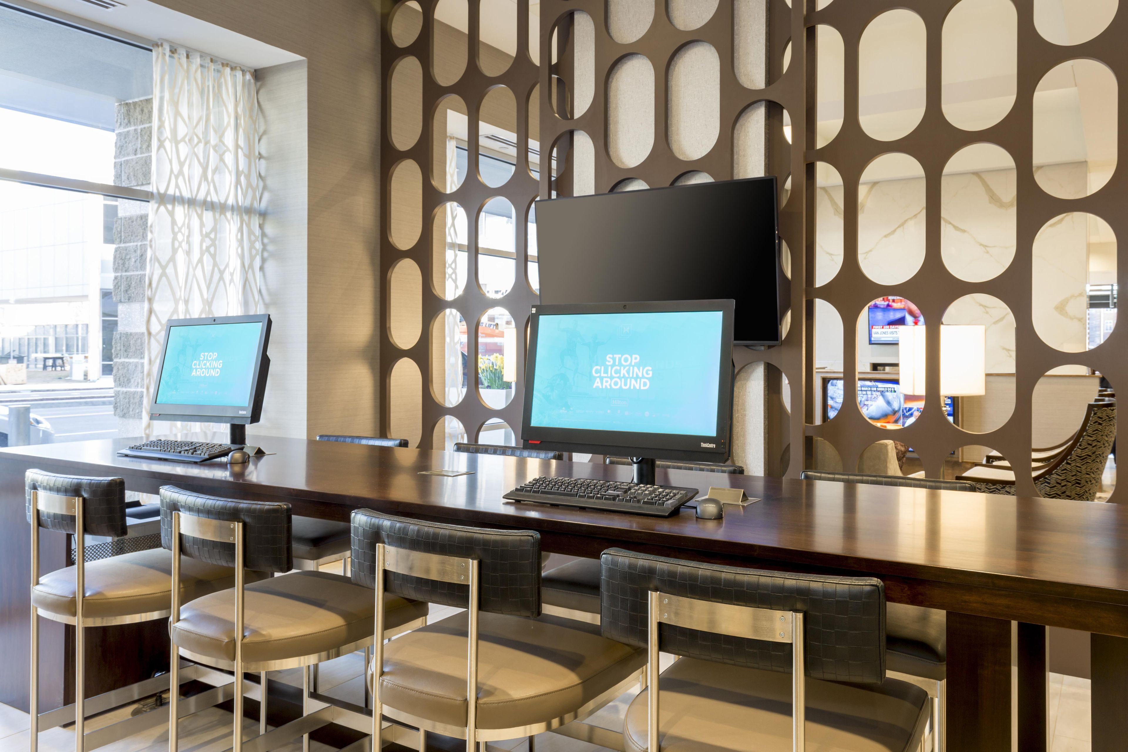 Un espace d’accueil d’hôtel comprenant deux postes de travail informatiques, un coin salon avec des fauteuils et un mur décoratif orné d’ouvertures circulaires.