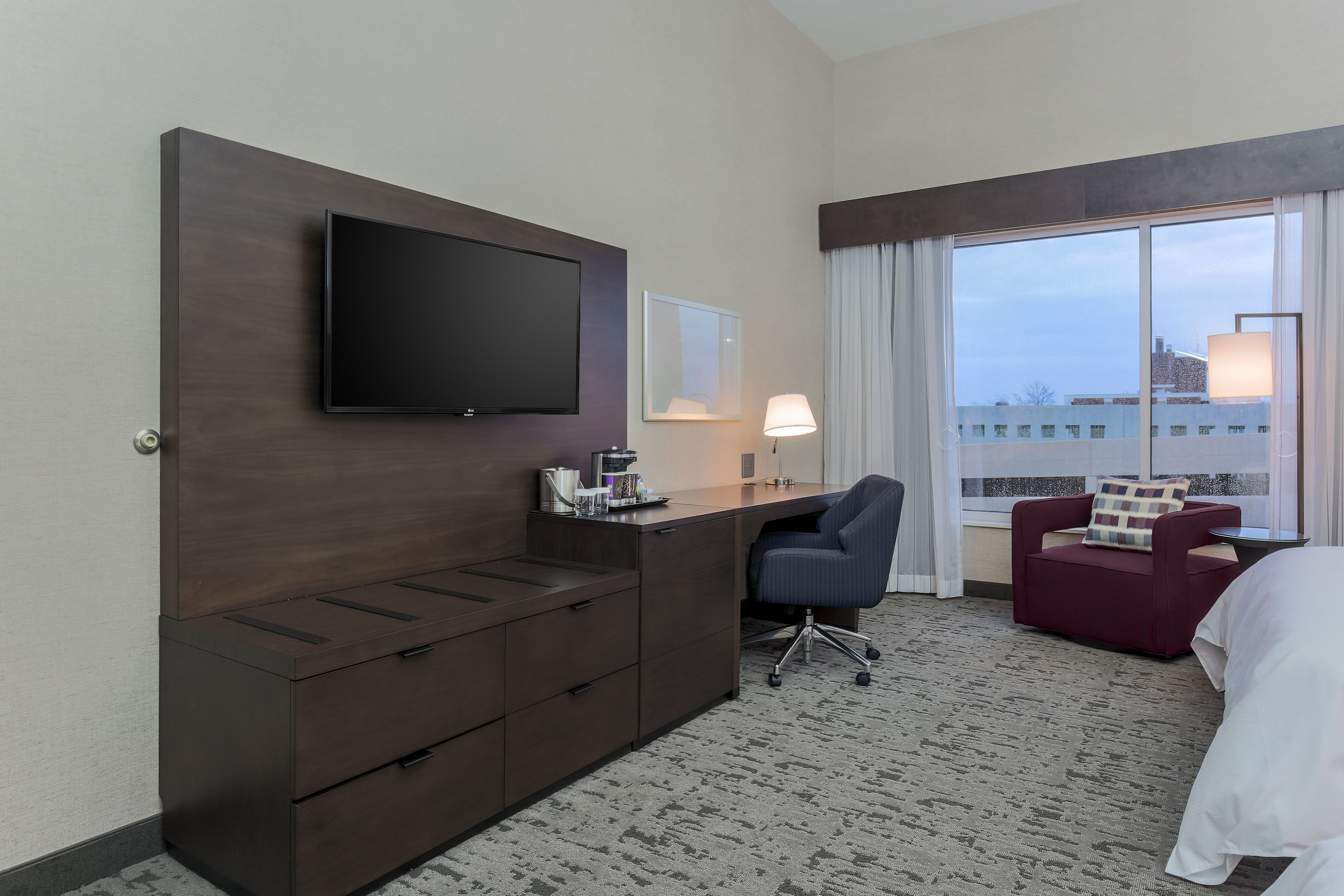 Une chambre d’hôtel dotée d’un téléviseur à écran plat fixé sur une commode en bois foncé, d’un bureau avec une lampe, d’un fauteuil rouge et d’une grande fenêtre donnant sur la ville.