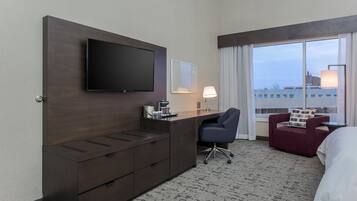 A hotel room with a flat-screen TV mounted on a dark wooden dresser, a desk with a lamp, a red armchair, and a large window offering a city view.