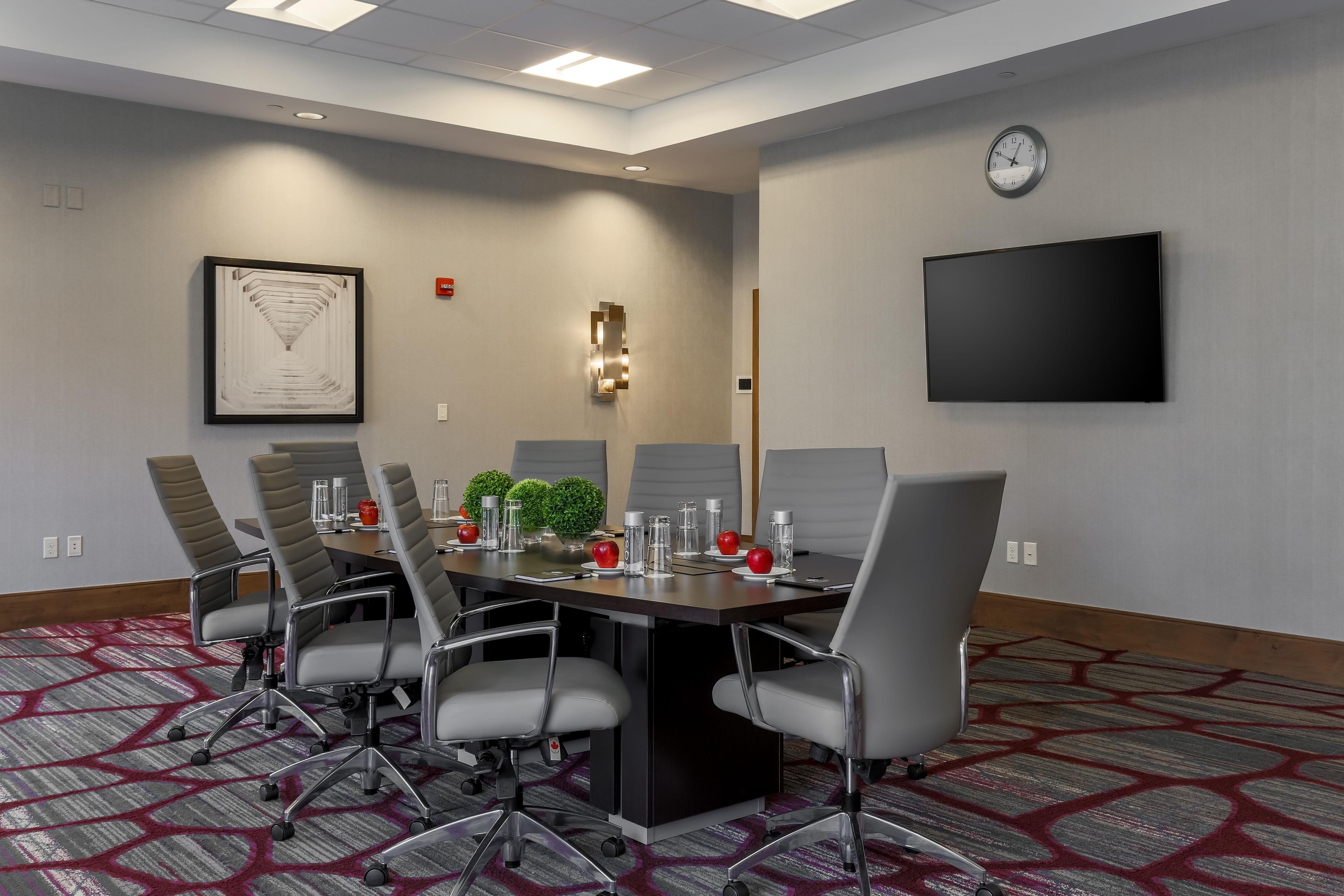 A conference room with a long table, six chairs, a clock, a framed artwork, and a flat-screen TV on the wall.